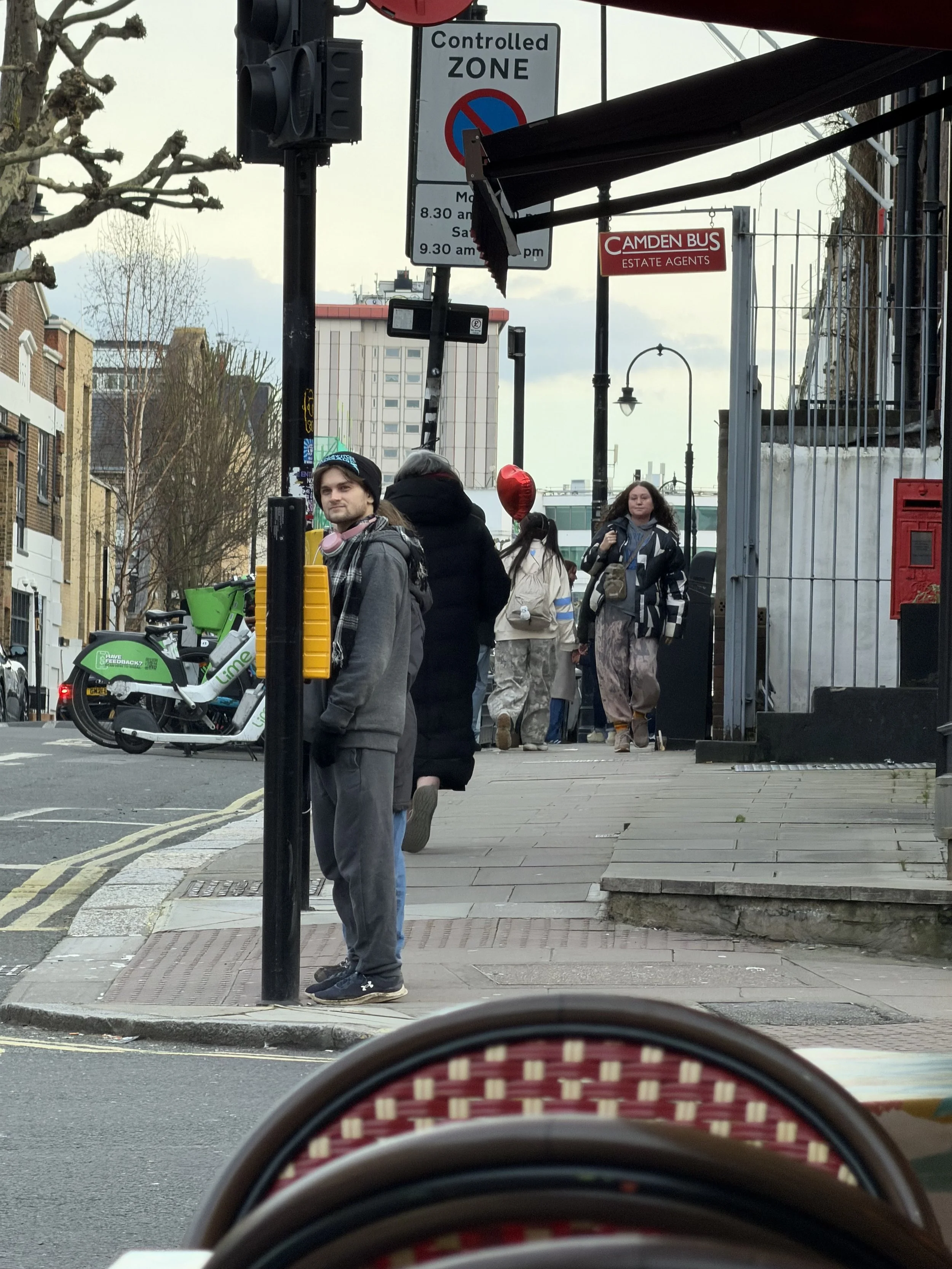 A busy city sidewalk with pedestrians, including a man in a hoodie and gray pants, standing at the curb. There are several people walking, some carrying bags, and a woman with wide pants and a backpack. A green and white bike-sharing scooter is parke