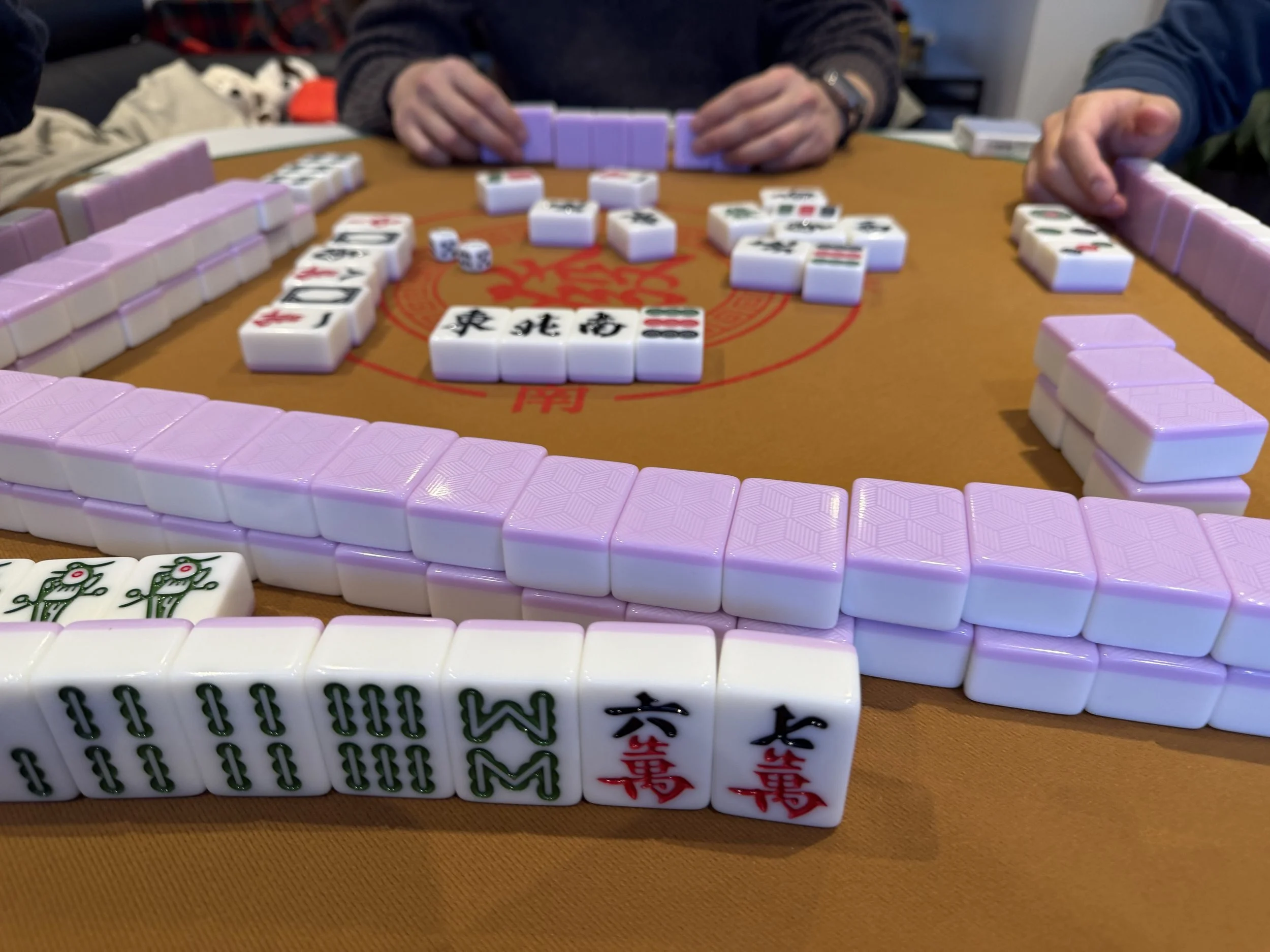 A group of people playing Mahjong with tiles on a brown table.