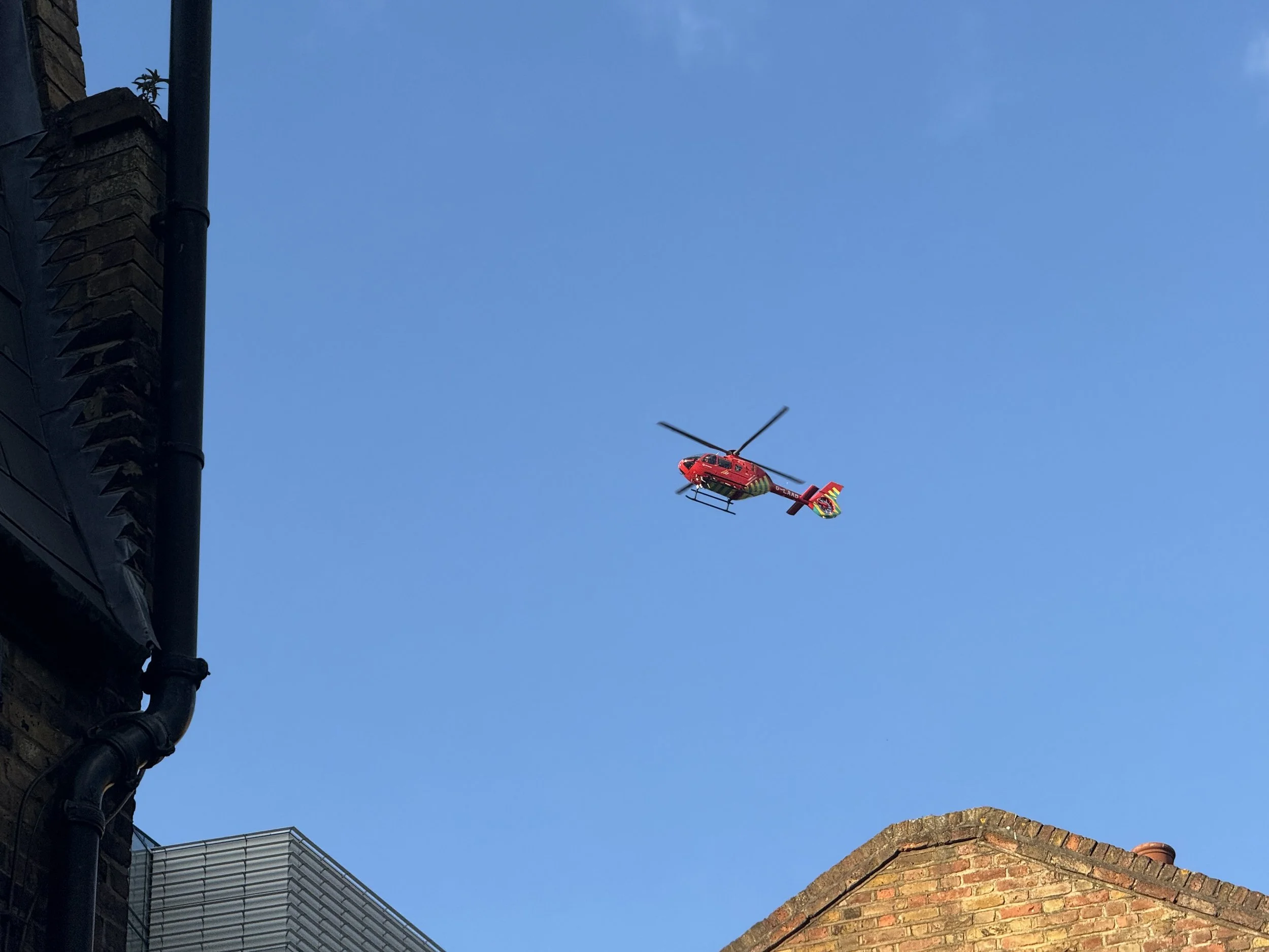 Red helicopter flying in clear blue sky with rooftops and building on the edges of the image.
