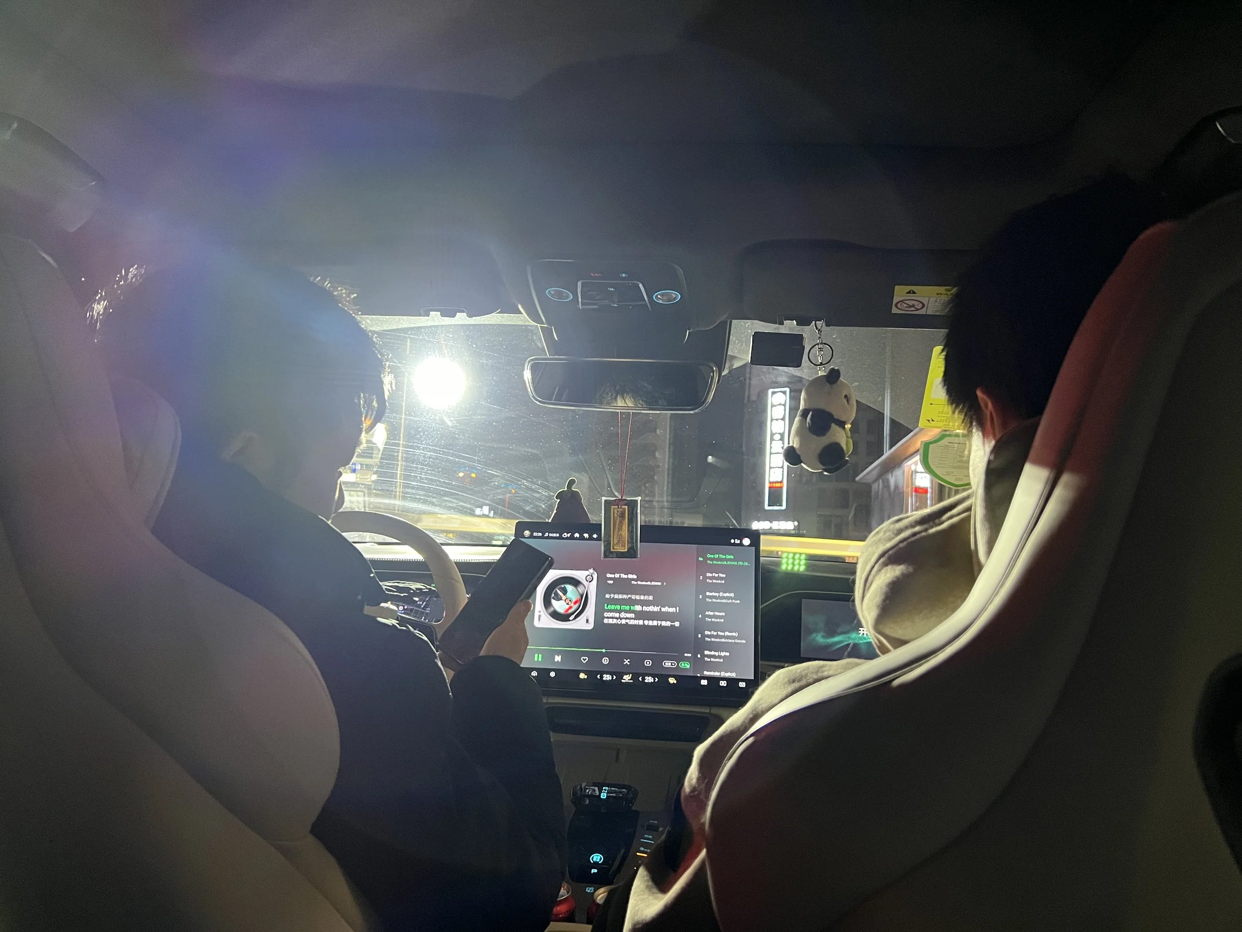 Inside a car at night, showing the back of the driver and passenger seats, someone holding a phone, a rearview mirror, a panda plush toy hanging from the ceiling, a dashboard with an electronic display, and a rearview mirror.