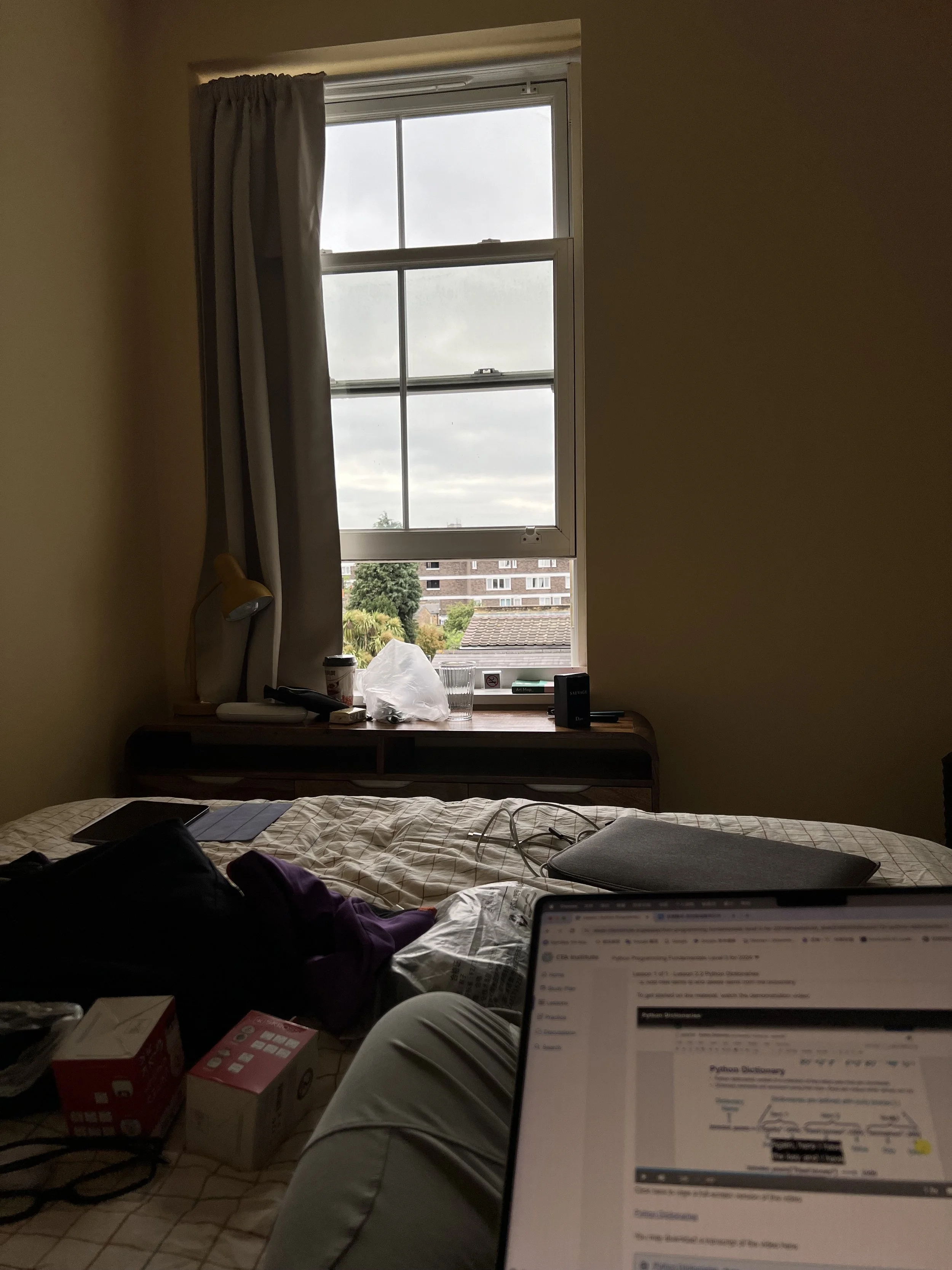 A view of a bedroom with an open window, showing residential buildings and trees outside. The window has a curtain on one side and a variety of items on the windowsill. The bed is in the foreground with electronic devices, clothing, and boxes on the 