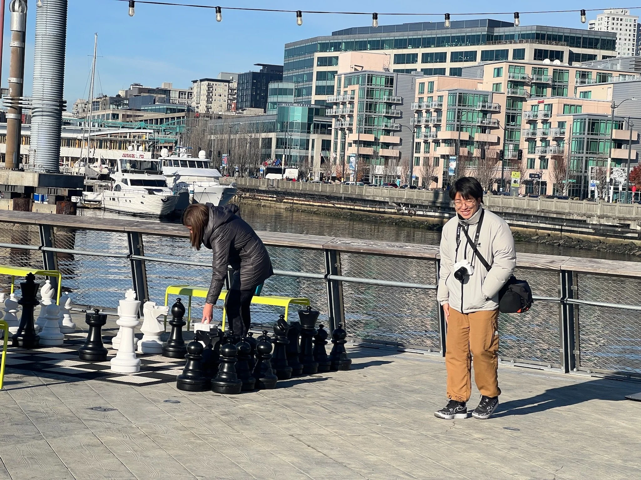 People playing chess on an outdoor waterfront promenade with boats docked nearby and modern city buildings in the background.