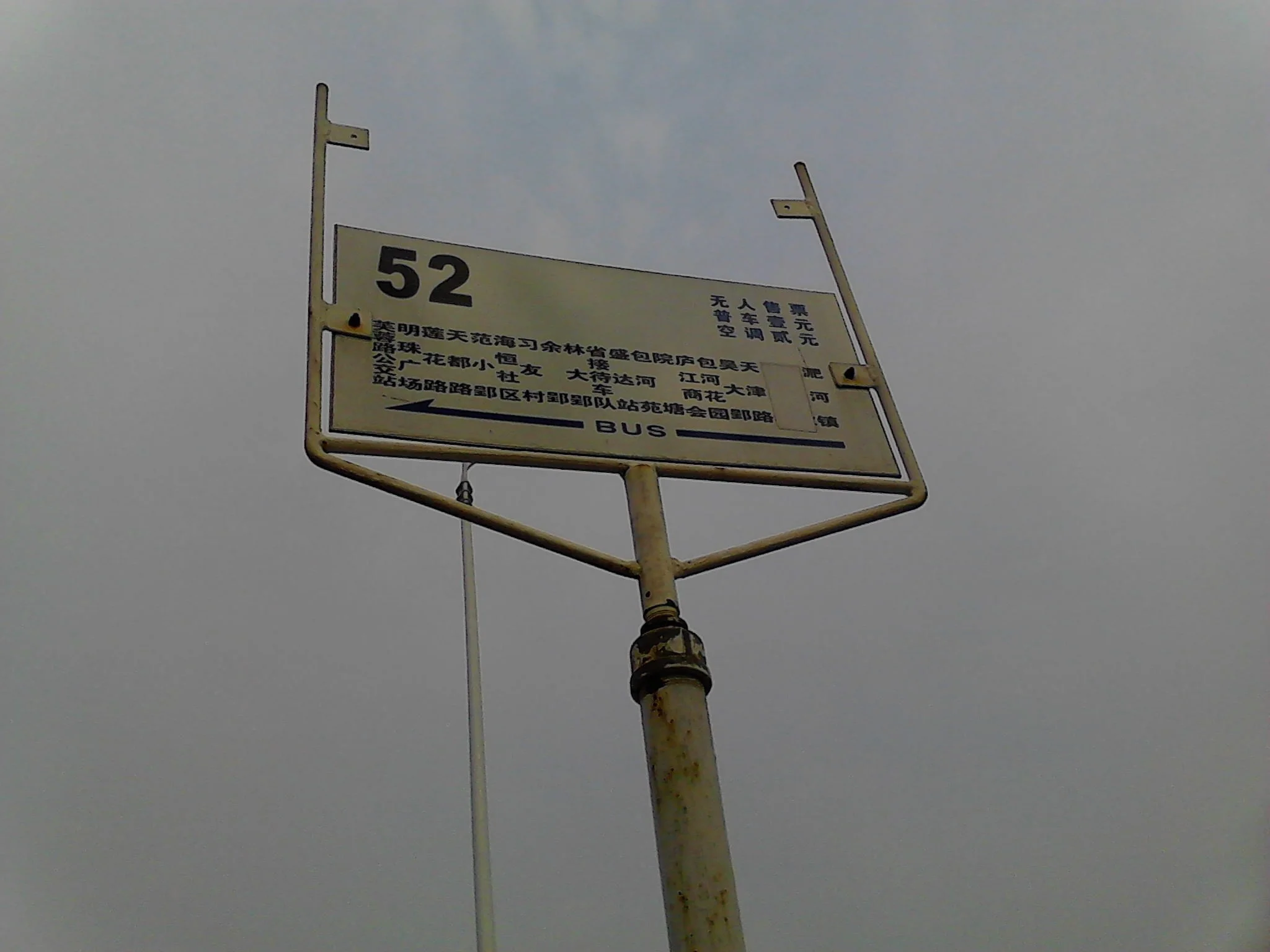 A bus stop sign with Chinese characters and the number 52, mounted on a yellow, rusted metal pole against a cloudy sky.