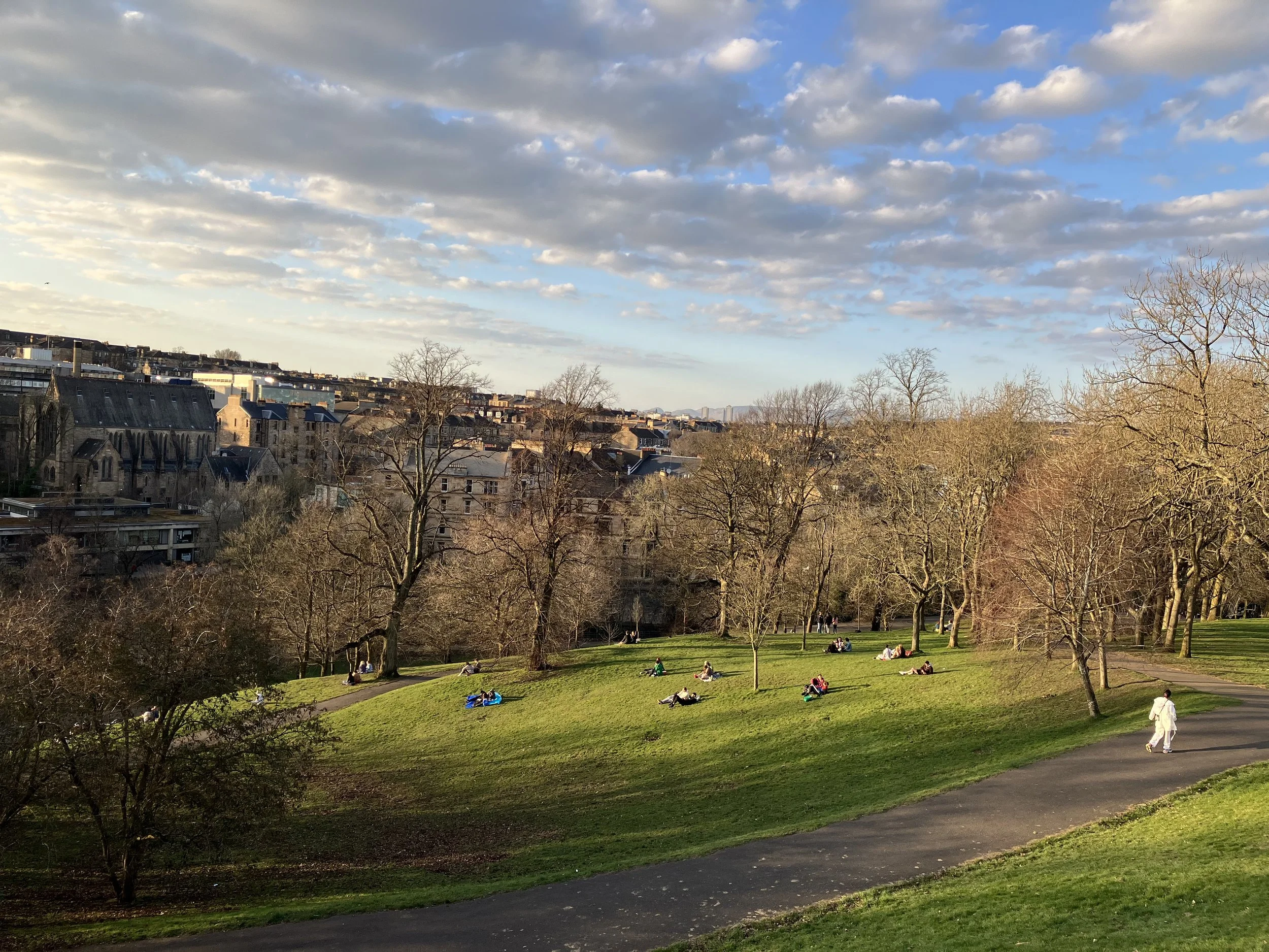 A park withpeople sitting and lying on the grass, trees without leaves, and city buildings in the background under a sky with scattered clouds.