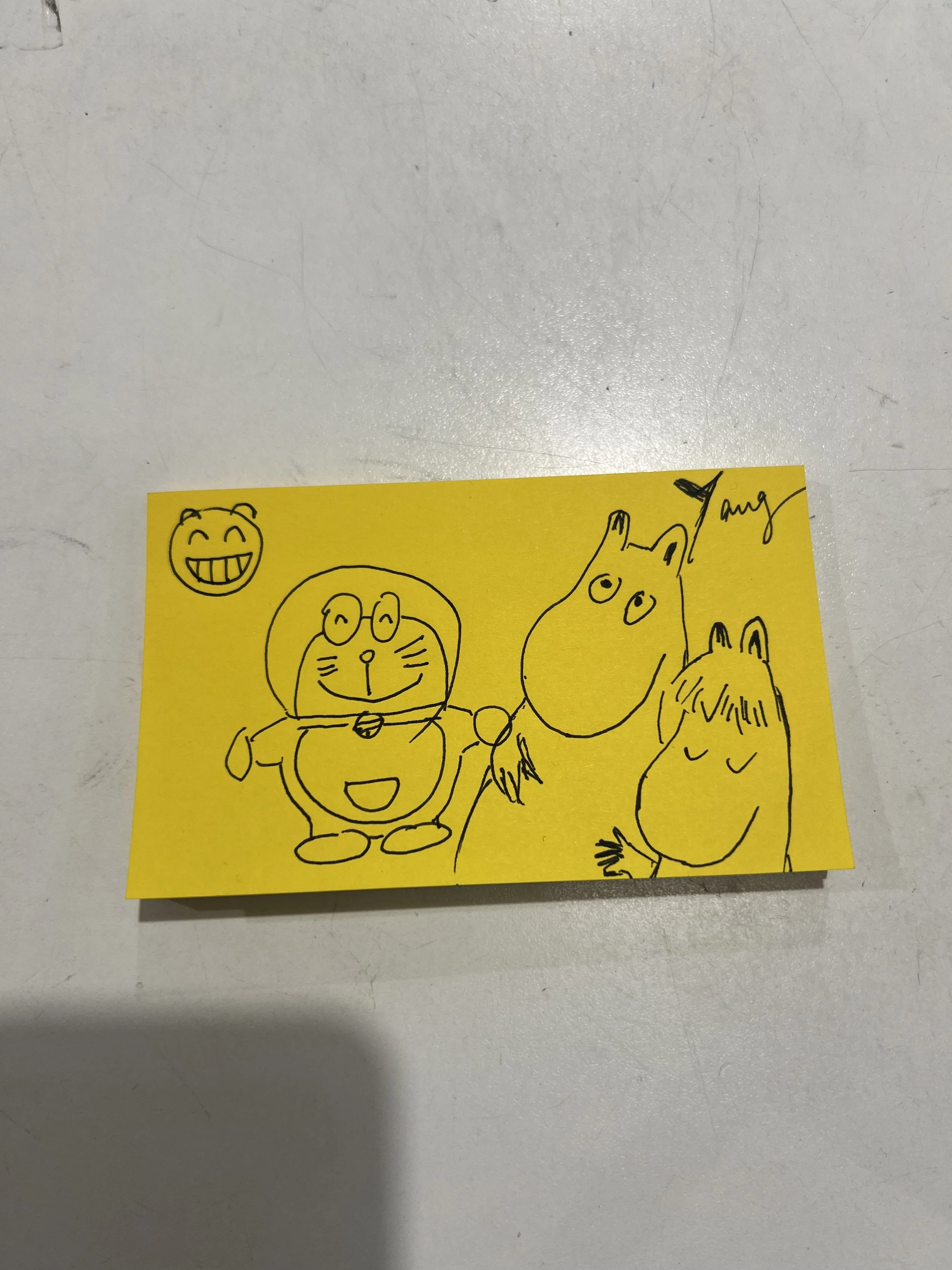 A yellow sticky note with black ink drawings of various cartoon characters, including a smiling face, a round-headed creature with glasses, a horse, and a person with long hair.