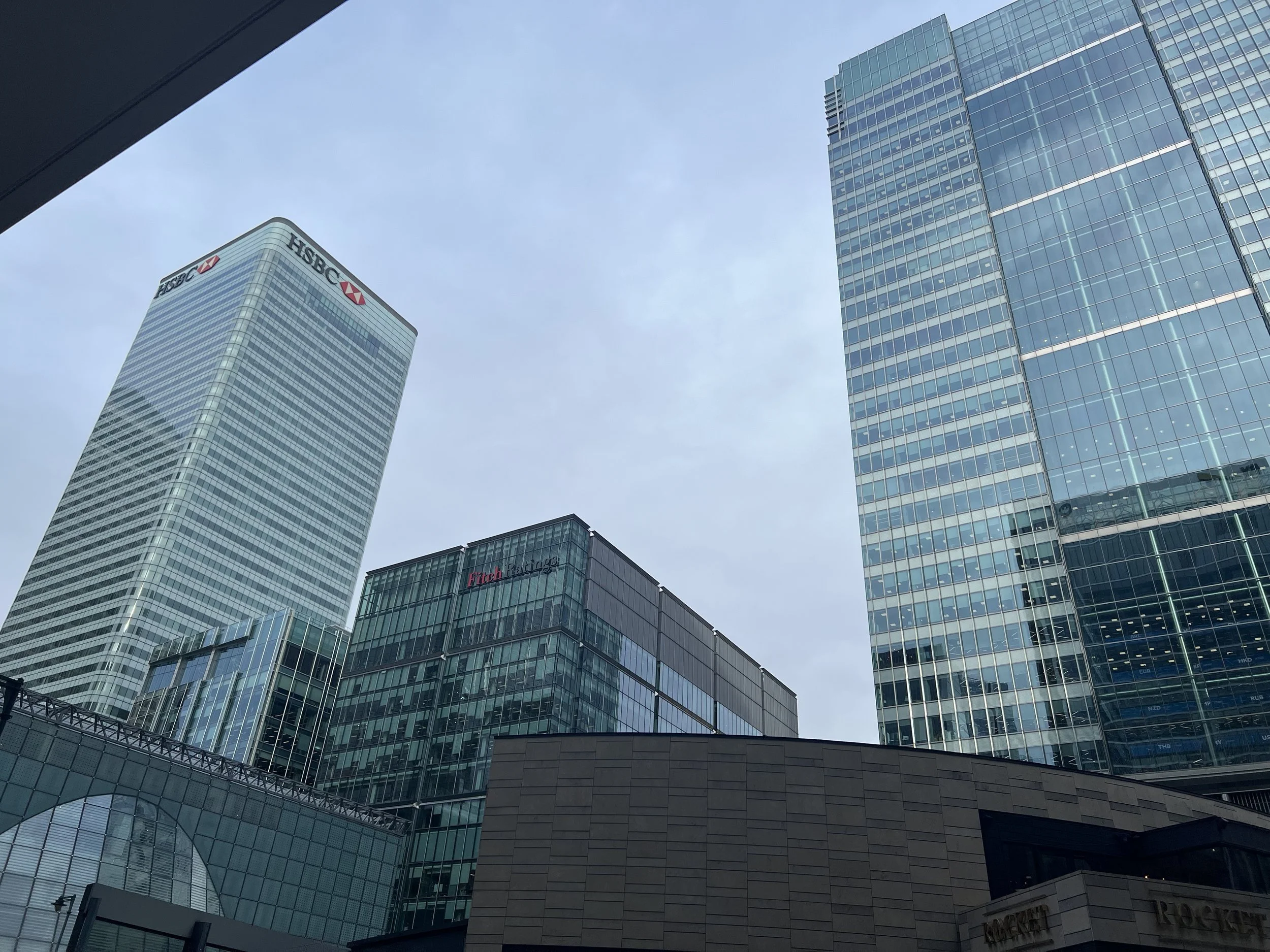 View of tall modern glass skyscrapers, including HSBC and Fitch Ratings buildings, with a blue sky in the background.
