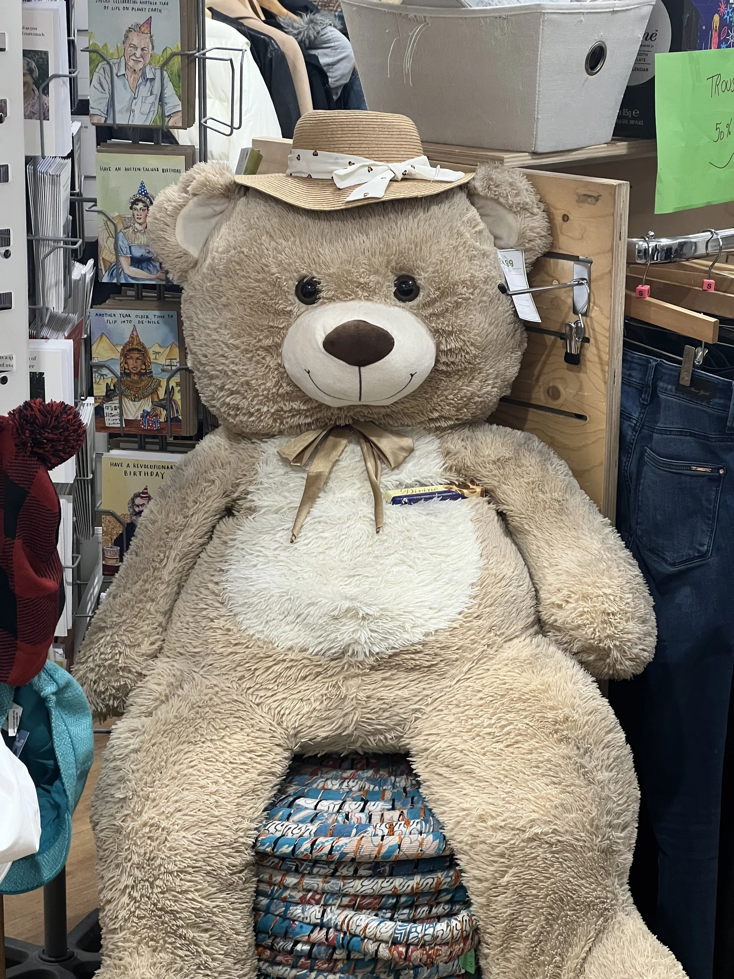 Large beige teddy bear with a white face and dark nose, wearing a straw hat with a white ribbon, sitting on a display shelf among greeting cards and other items in a store.