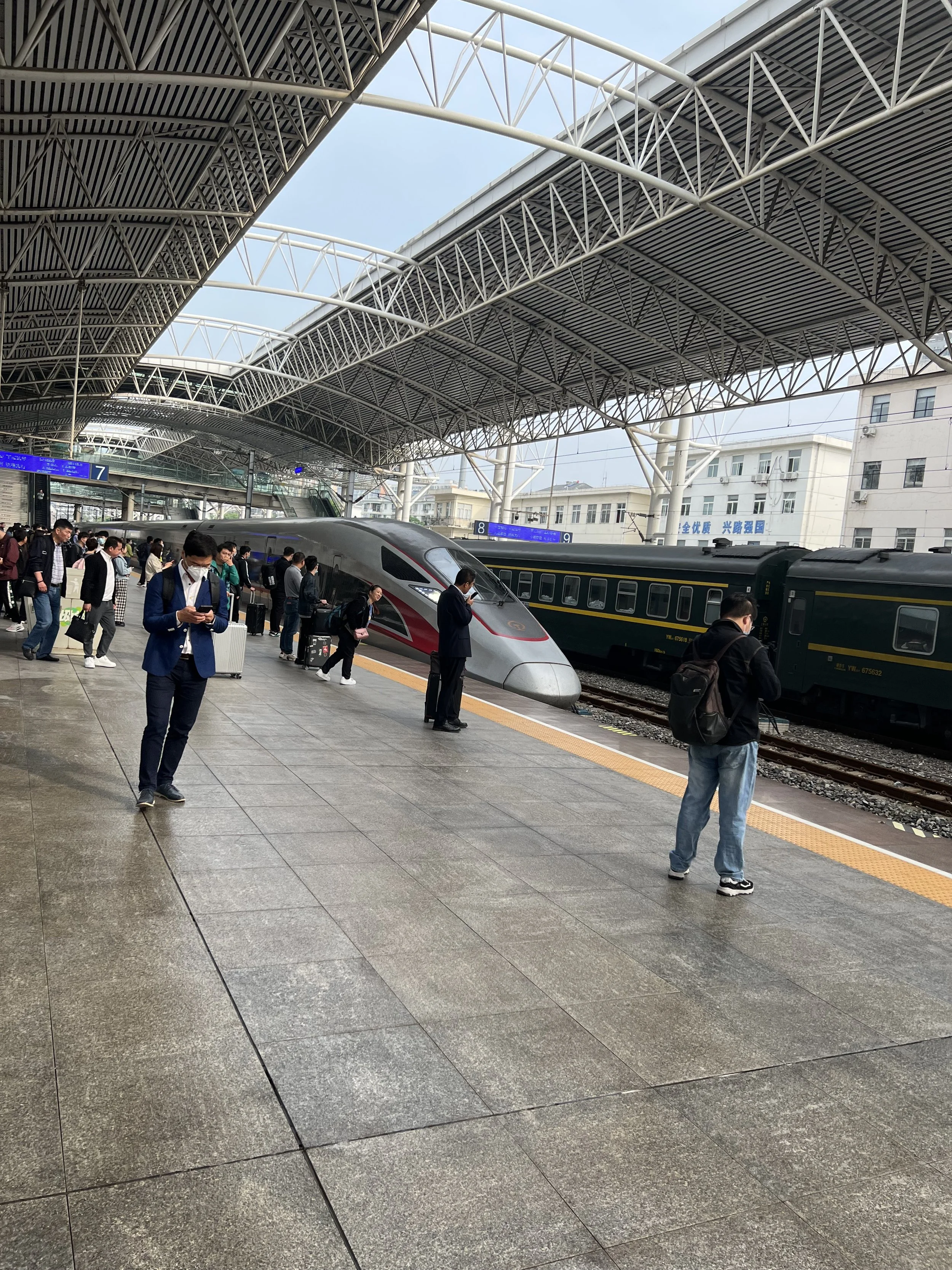 People boarding and waiting for trains at a modern train station with a covered roof and urban buildings in the background.