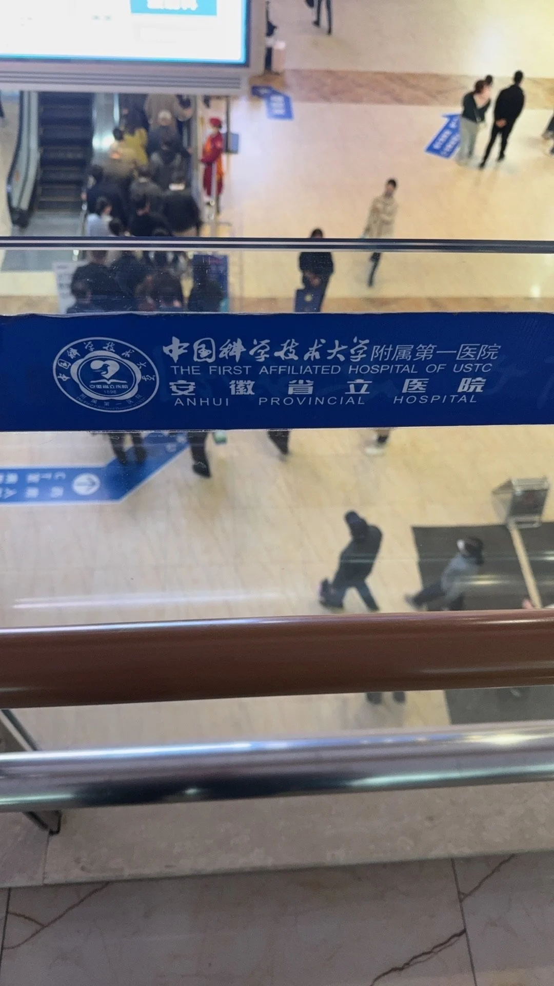 View from inside a building looking down on a busy lobby with people walking and sitting, seen through a glass barrier with a blue label from Anhui Provincial Hospital, affiliated with USTC in China.