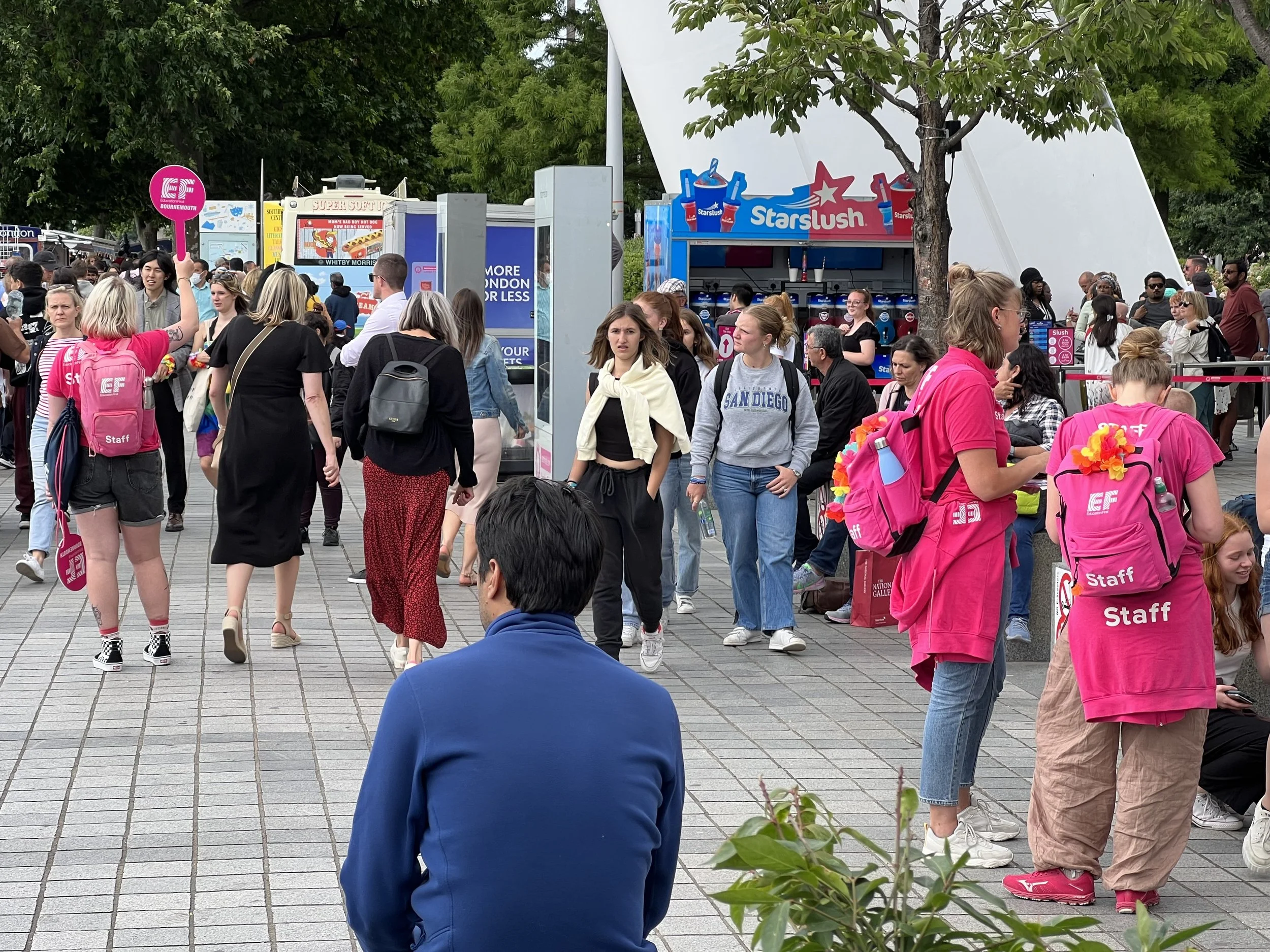 Crowd of people, some wearing backpacks with staff badges, gathered at an outdoor event with food stalls and trees in the background.