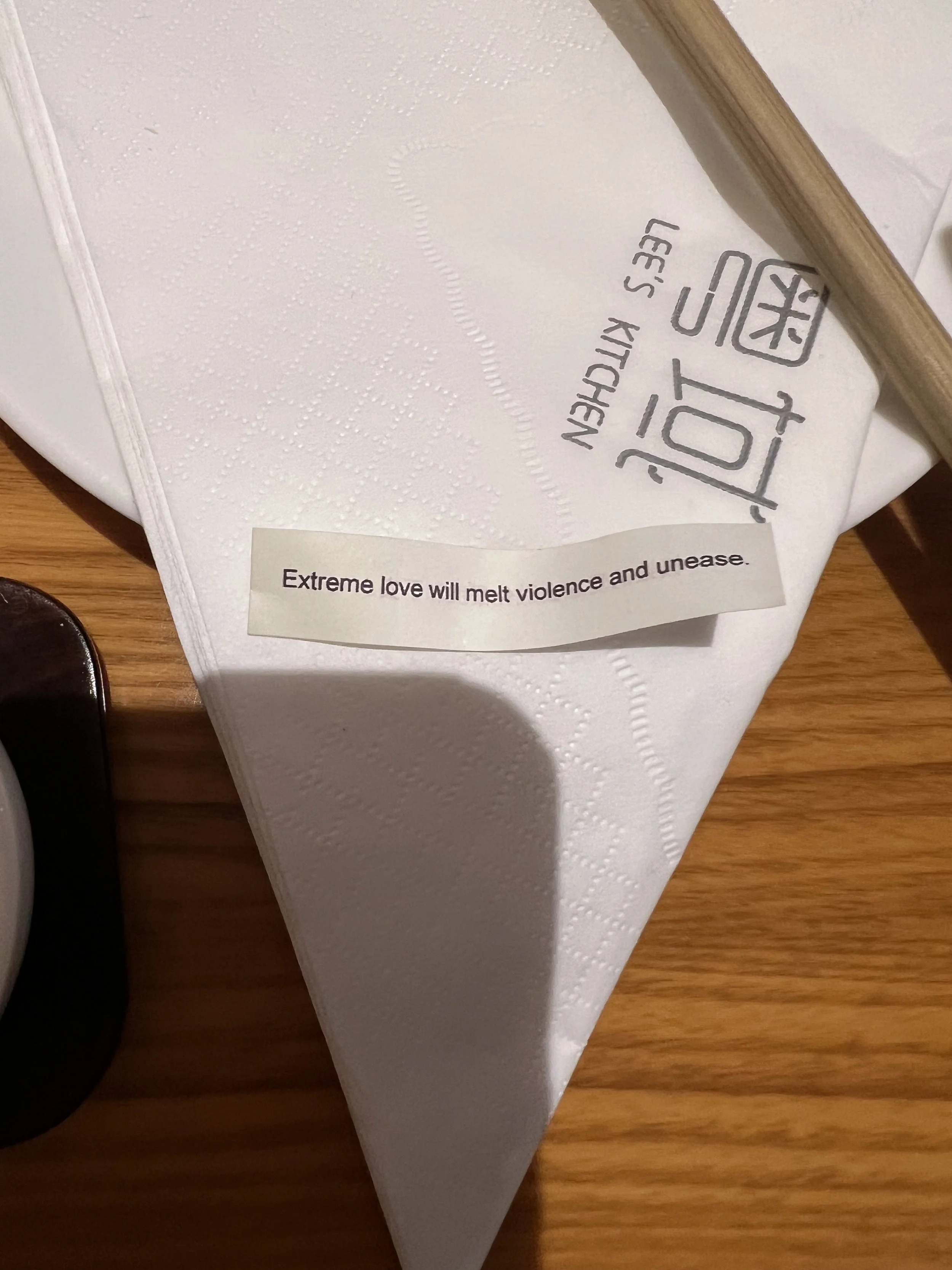 White paper napkin with ink and embossed patterns, placed on a wooden table. Partially covered by a wooden-handled utensil. A label with text that reads 'Extreme love will melt violence and unease.' is attached to the napkin.