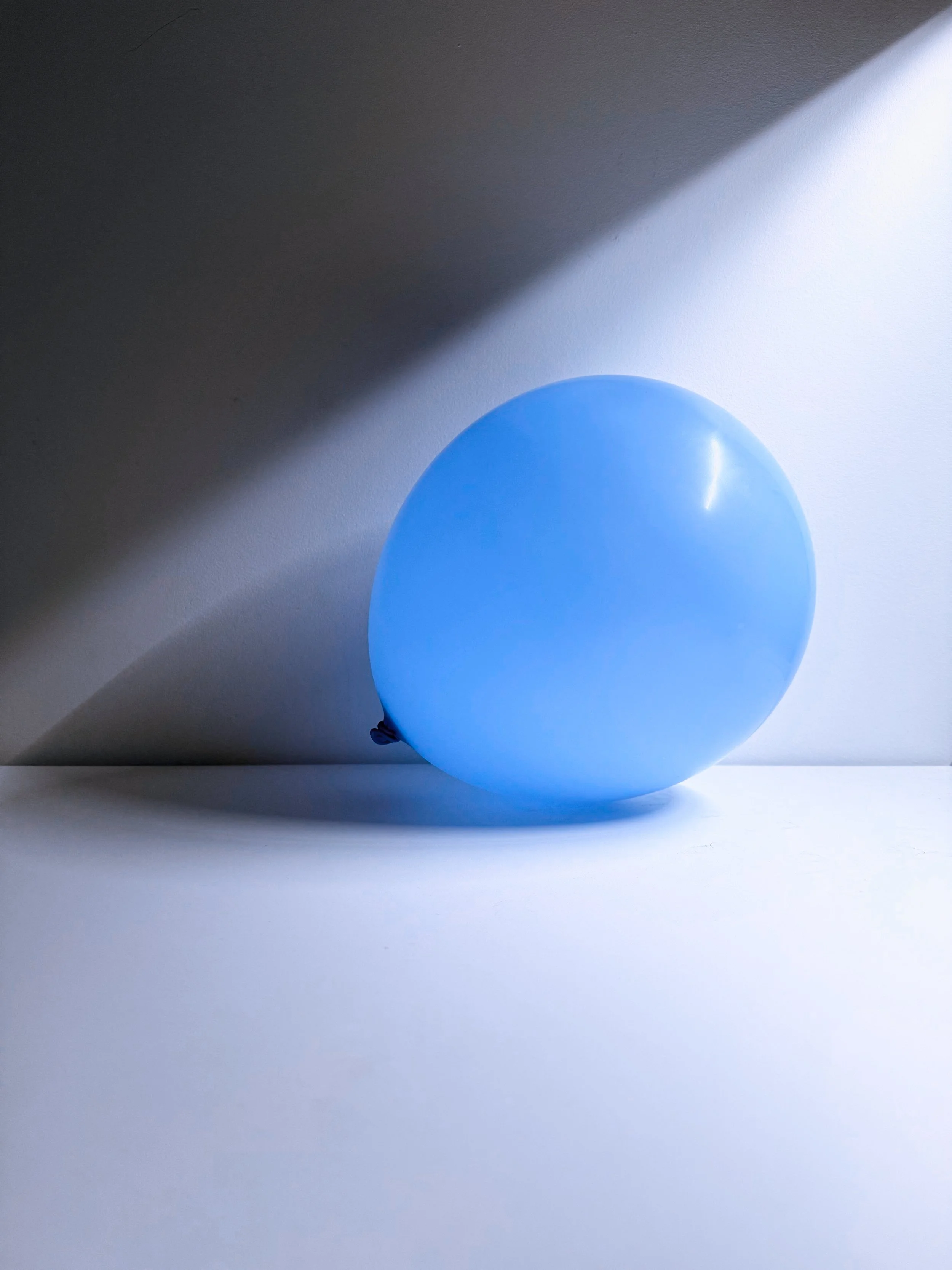 Blue balloon on a white surface with a shadow cast by the balloon and a shadowed corner of the image.