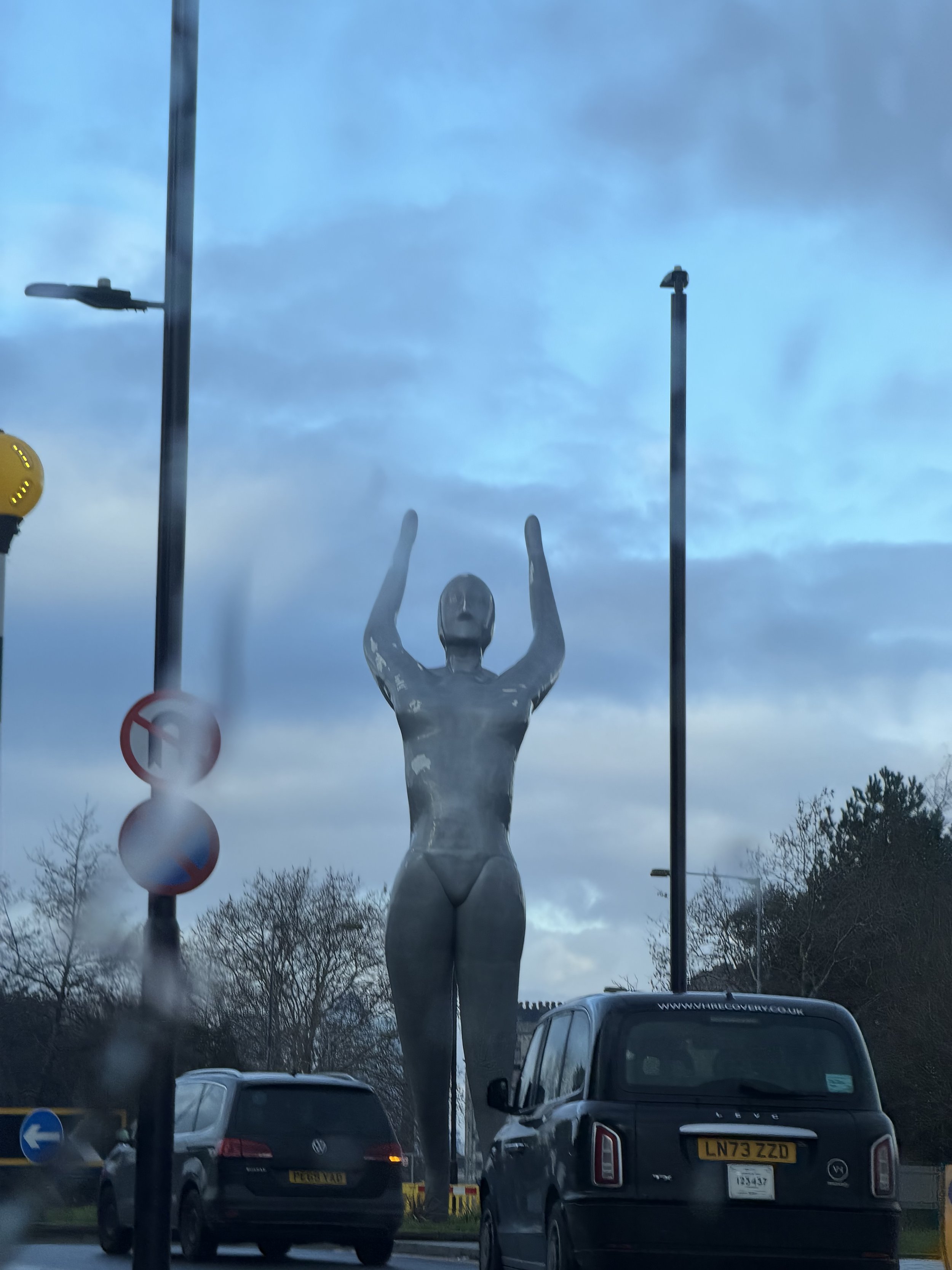 A street scene with cars parked in front of a large statue of a woman with arms raised, flanked by streetlights and traffic signs, under a cloudy sky.