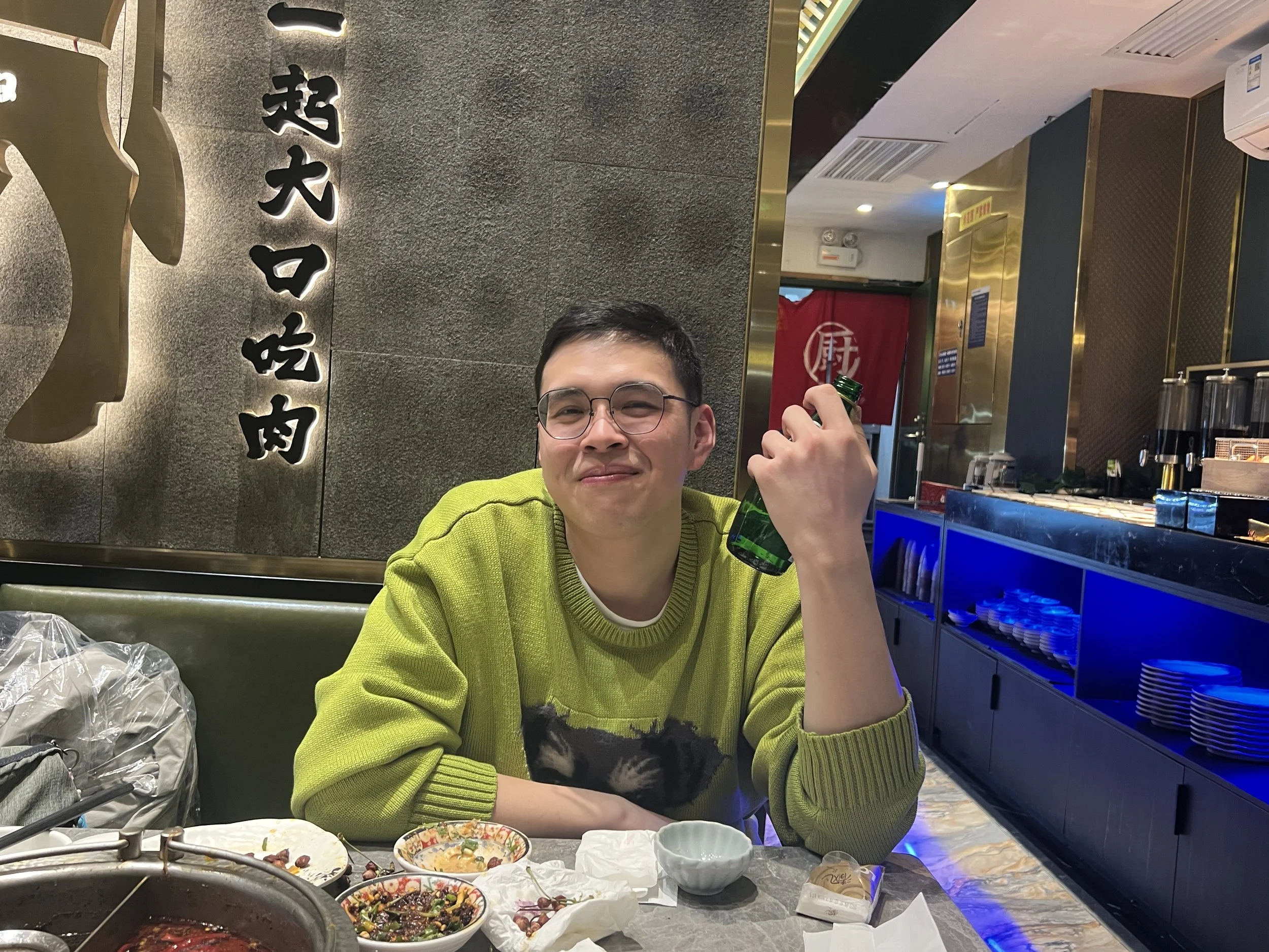 A young man with glasses wearing a lime green sweater smiling and holding a green beer bottle at a restaurant. There are bowls of food on the table in front of him, and a gray textured wall with Chinese characters illuminated behind him. Part of the 