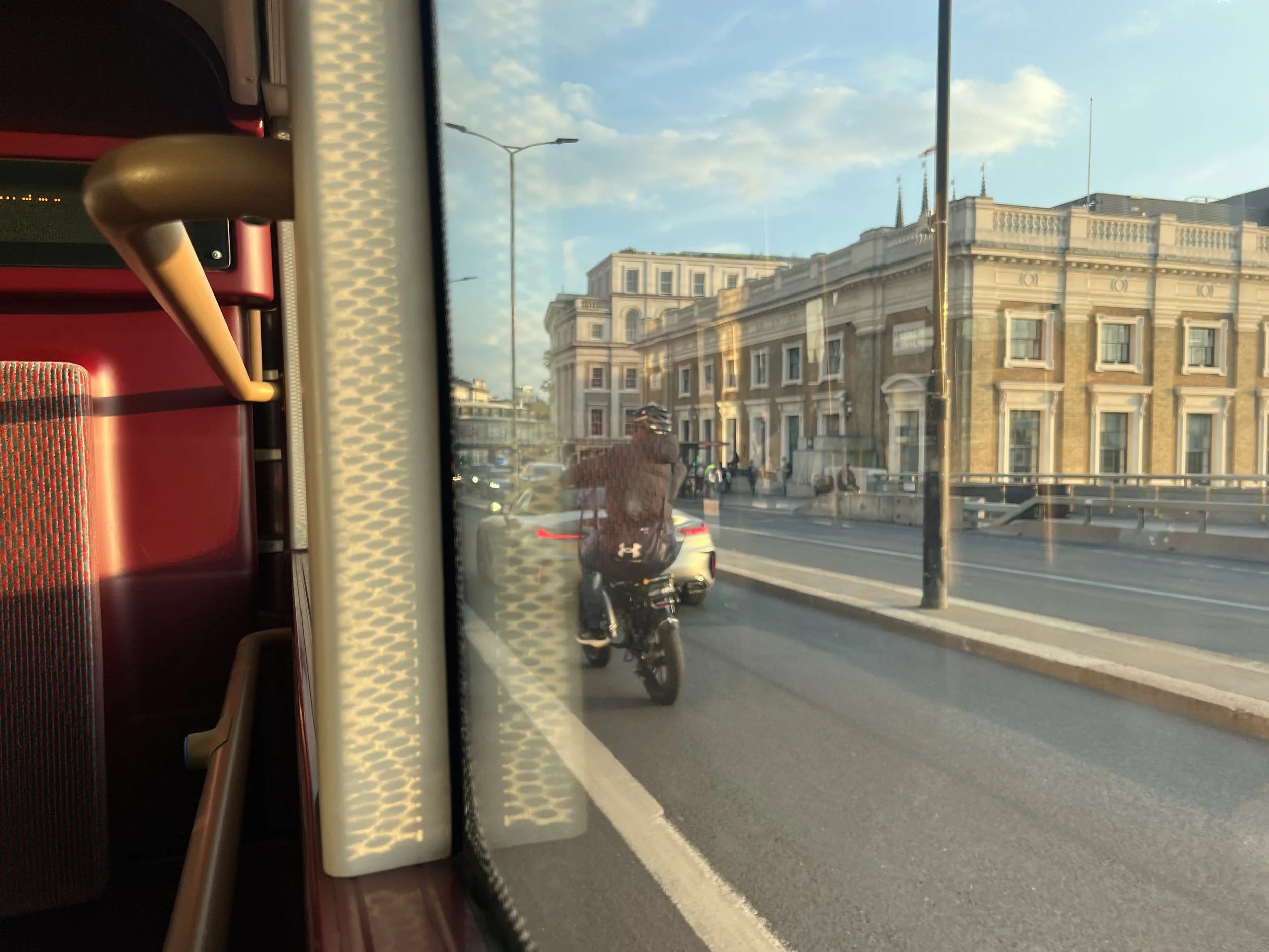 View from inside a bus showing a city street with a historic building, cars, and a lamppost outside.