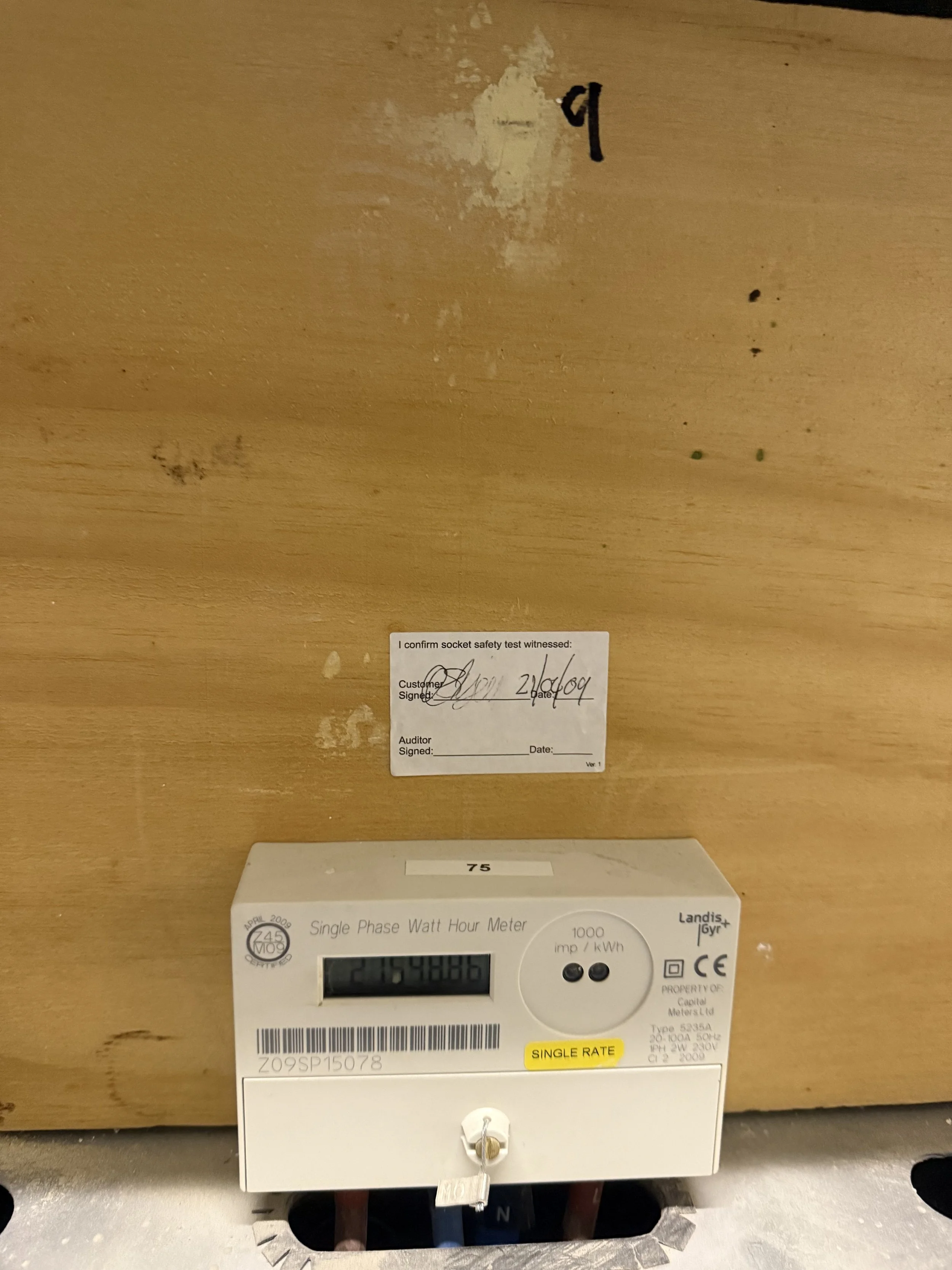 An electrical meter and an energy testing device mounted on a wooden surface, with a sticker showing the customer signing and the date, and a label indicating single rate electricity usage.