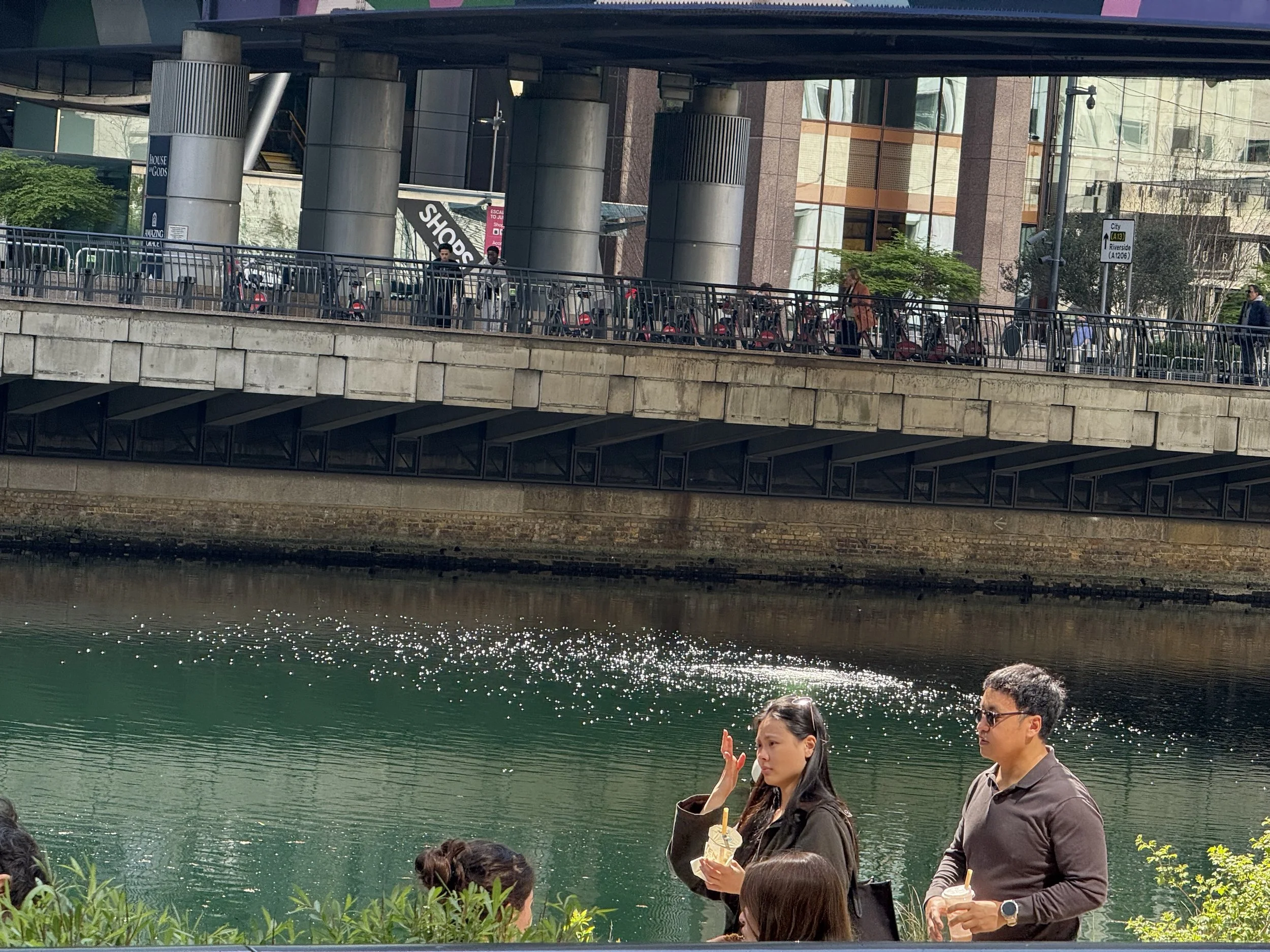 People standing near a riverbank with a building and a bridge above in the background.