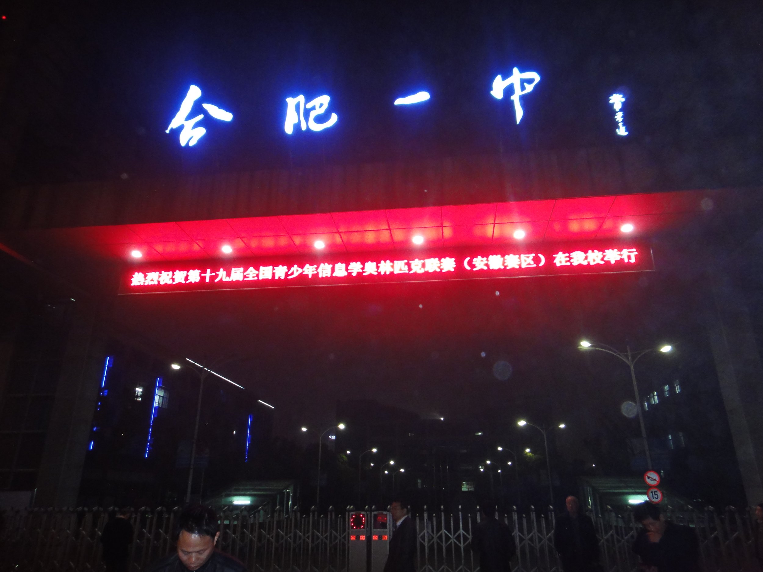 Night view of a building with bright blue lit Chinese characters and red neon signs, and a section of a toll gate or entrance barrier with people nearby.