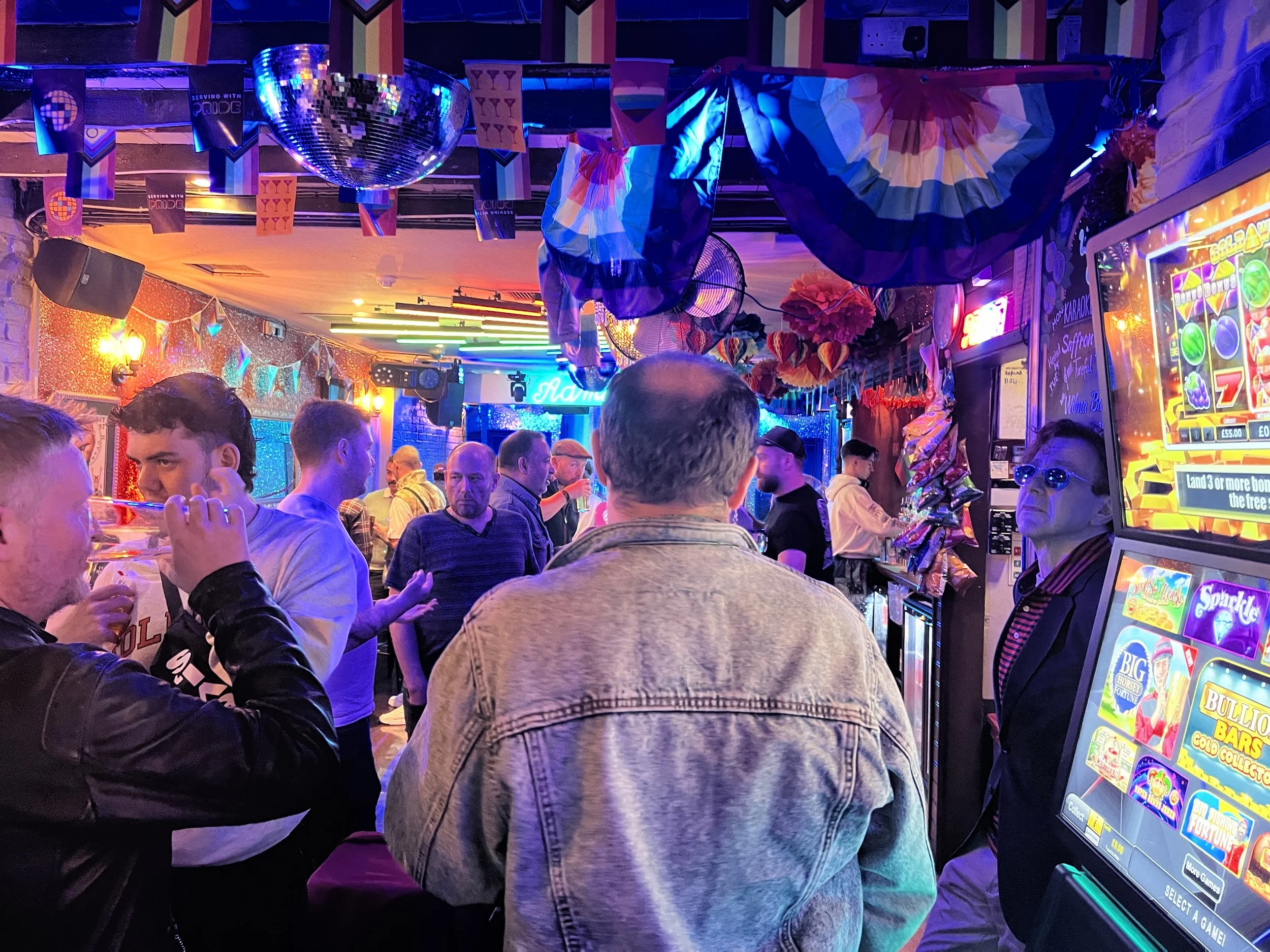Indoor scene at a bar or arcade with people socializing, colorful neon and disco lights, pride flags hanging from the ceiling, and gaming or slot machines to the right.