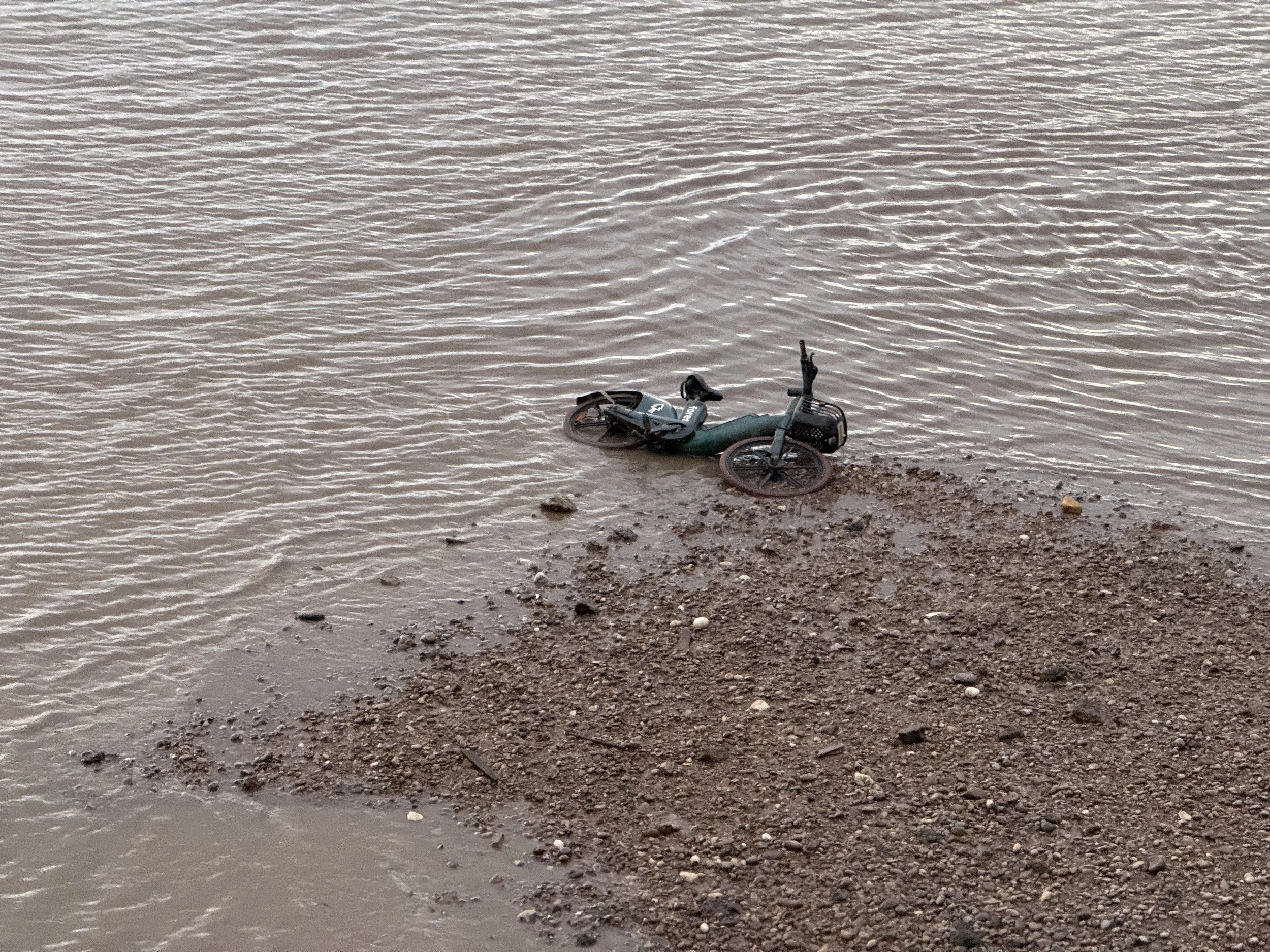 A motorcycle partially submerged and lying on its side on a muddy shoreline near a body of water.