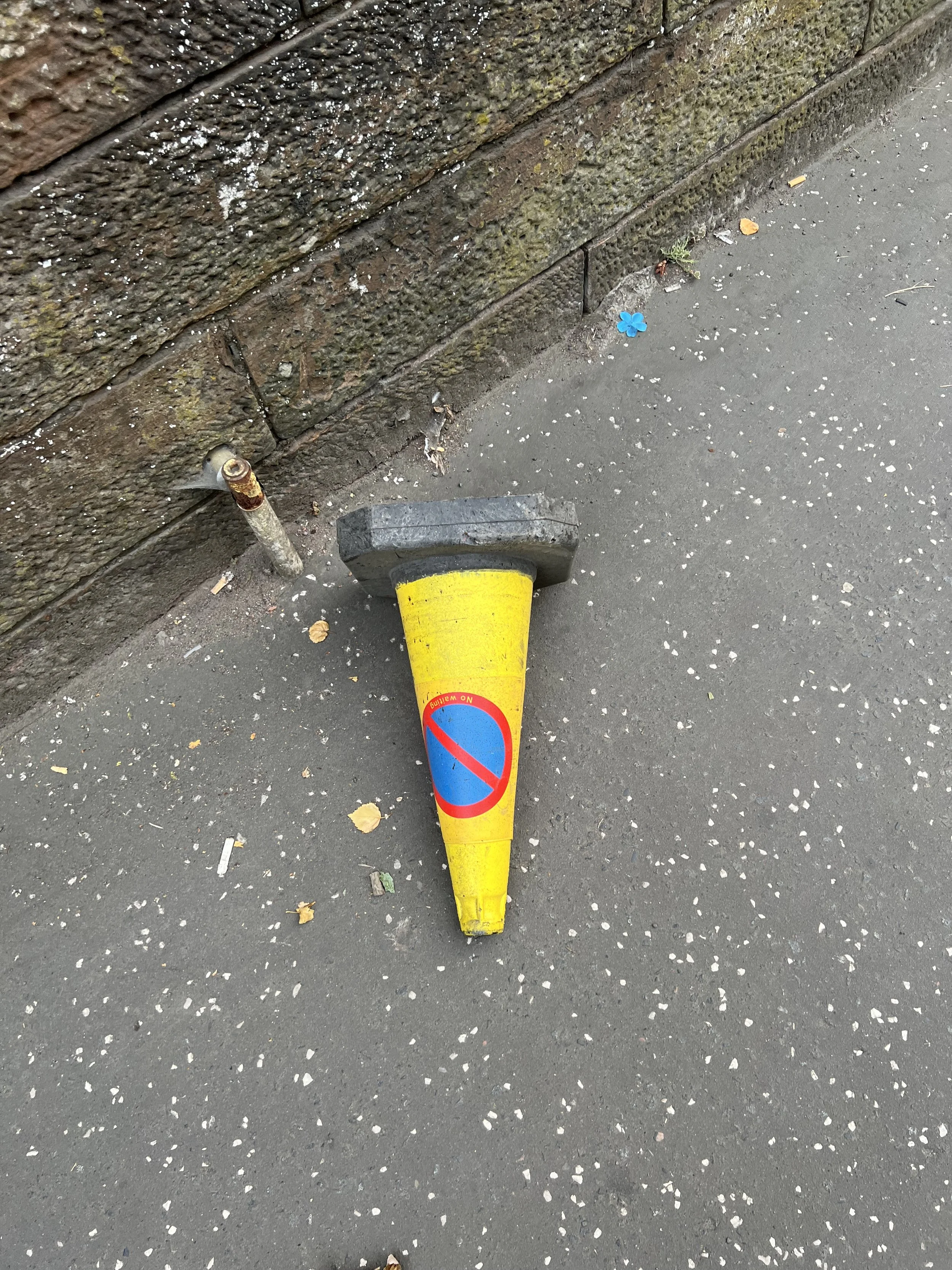 A yellow traffic cone with a no parking sign lying on its side on the sidewalk near a brick wall.