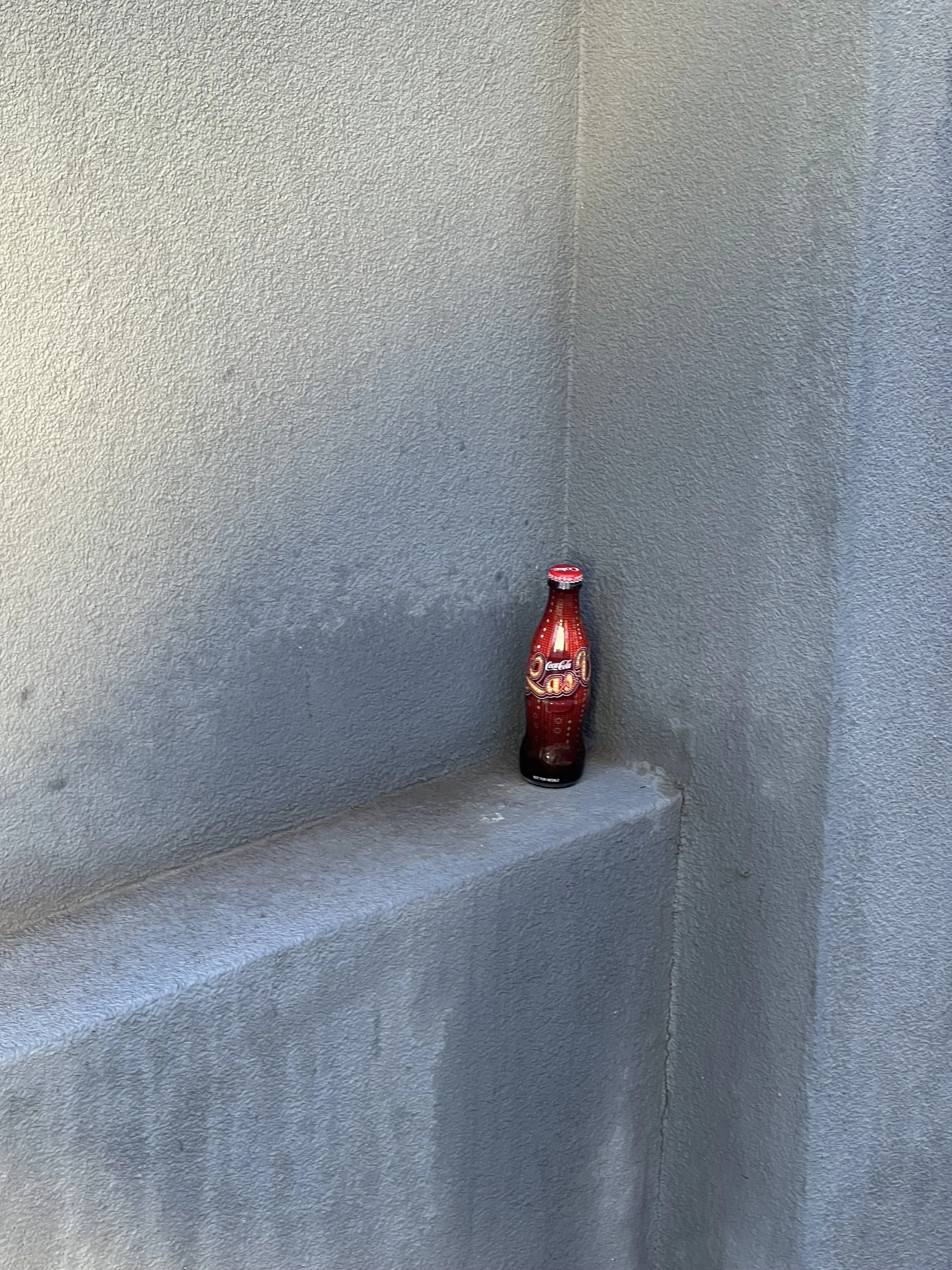 A red Coca-Cola bottle on a corner ledge against a textured gray wall.