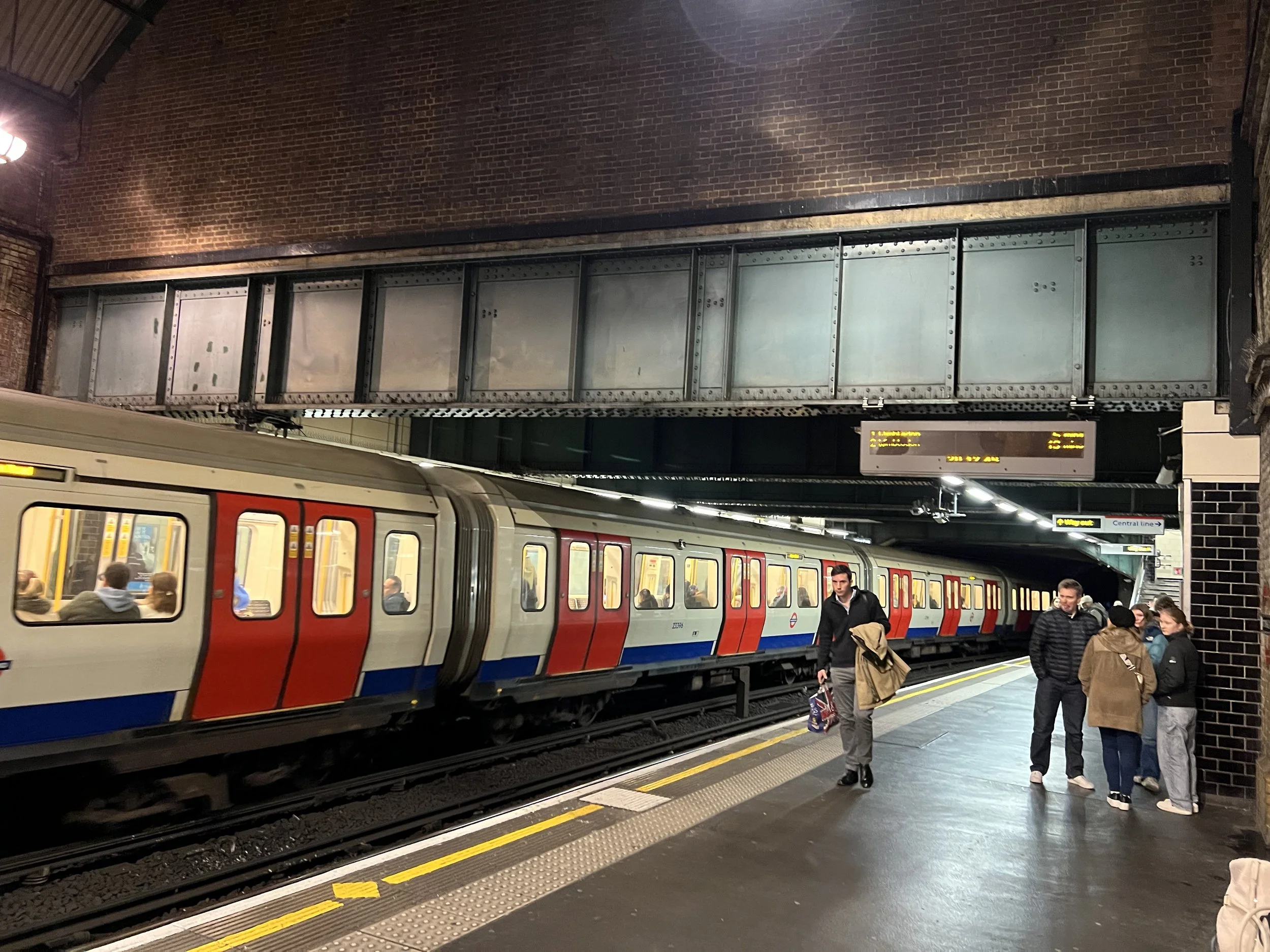 A busy underground train station with passengers waiting on the platform. A red, white, and blue London Underground train is at the station, with people entering and exiting. The background shows a brick wall and digital signs displaying travel infor