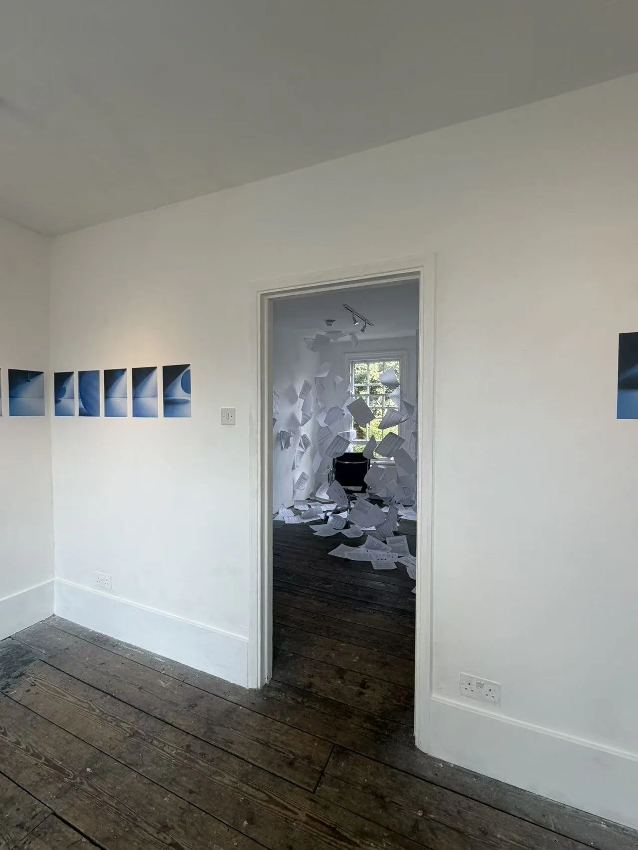 A room with white walls and wooden flooring. A doorway leads to another room with papers flying or falling through the air. The second room has a large window letting in natural light.