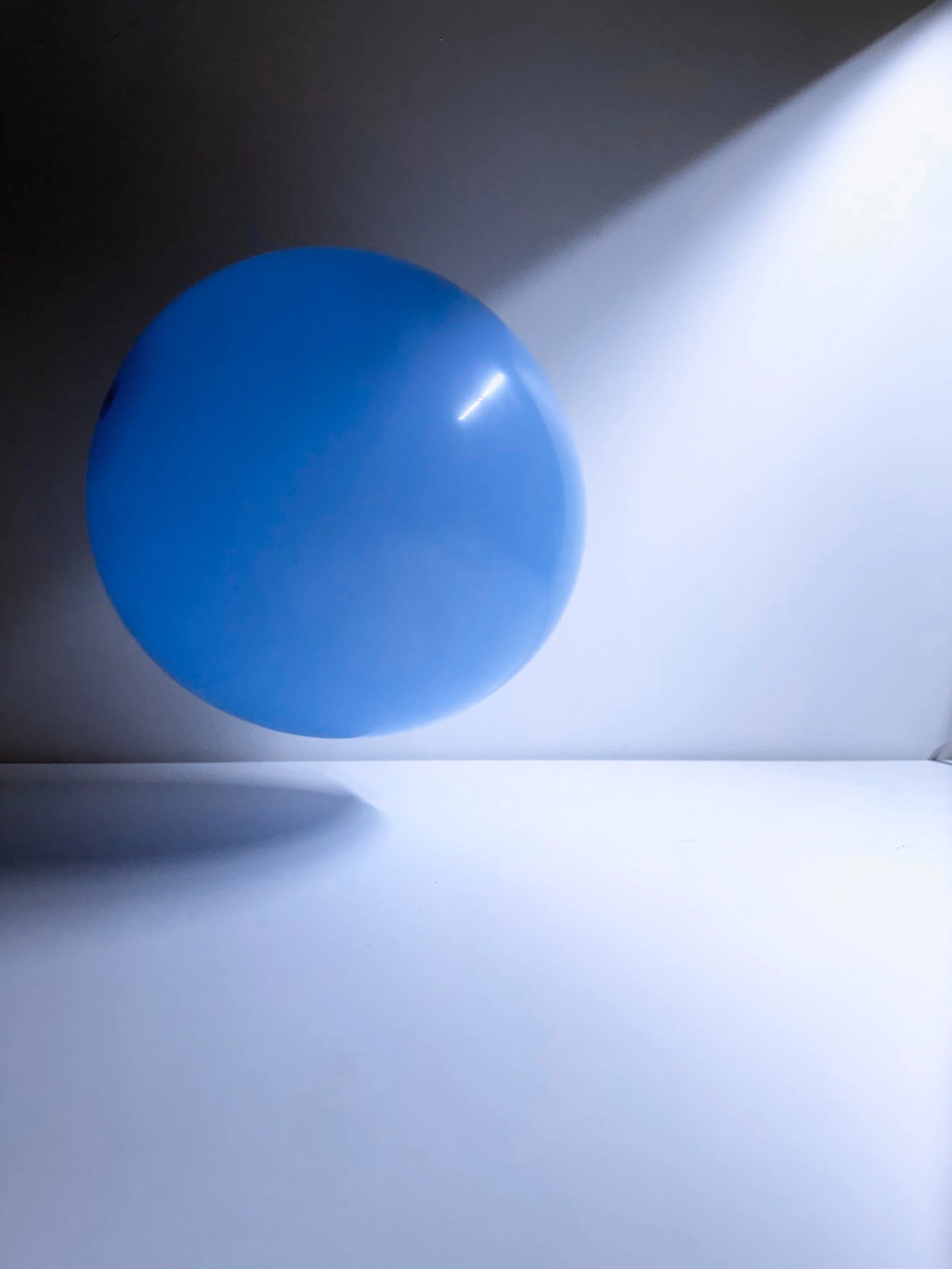 A blue balloon resting on a white surface with a shadow underneath it, cast by overhead lighting, and a bright light source illuminating the scene from the top right corner.