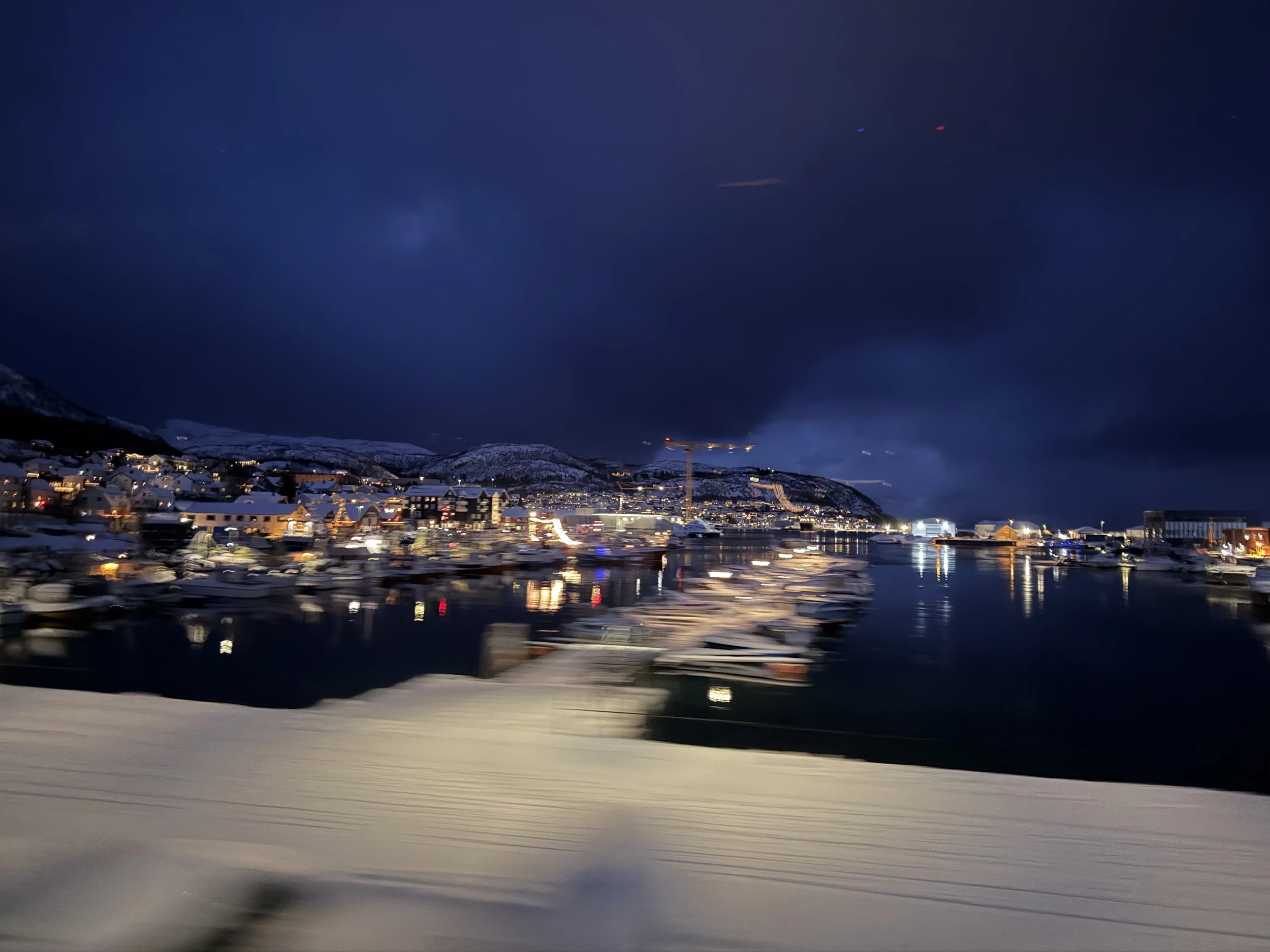 Night view of a seaside town with snow-covered buildings, boats docked in the harbor, and mountains in the background under a cloudy dark sky.