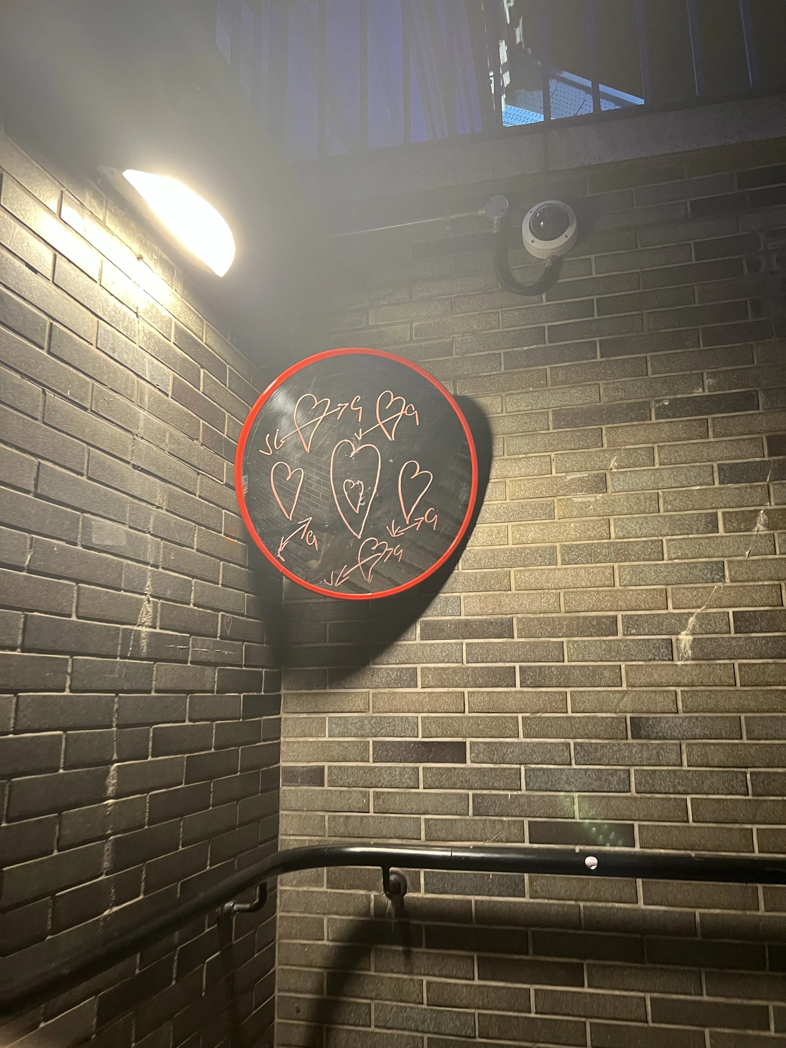 A brick wall with a white security camera mounted underneath a bright wall light. A black handrail is visible at the bottom of the image, and a black circular sign with pink hearts and handwritten-style writing is also present.