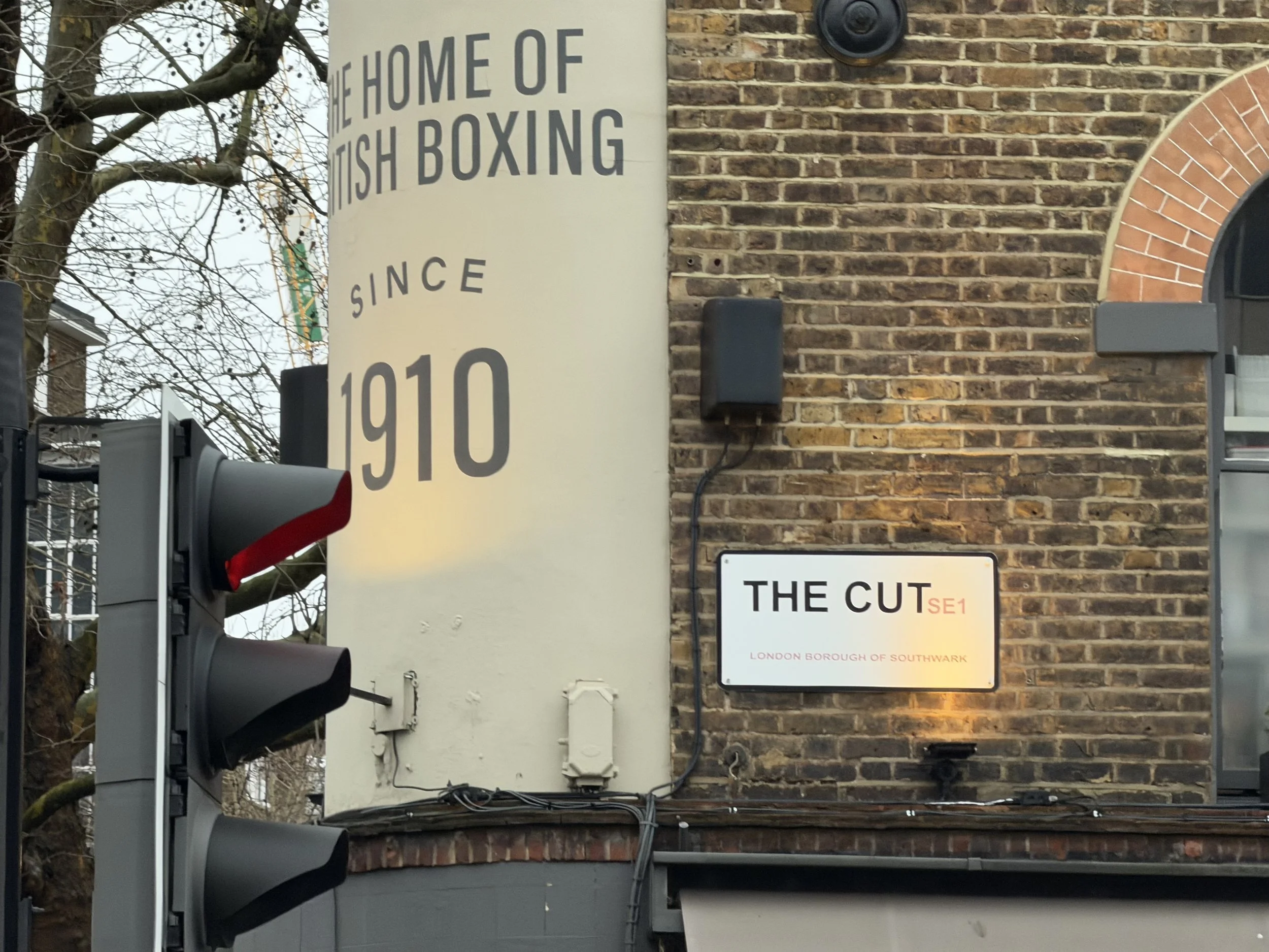 Street scene showing a sign reading 'The Home of British Boxing Since 1910' on a brick building wall, with traffic lights and some leafless trees in the background.
