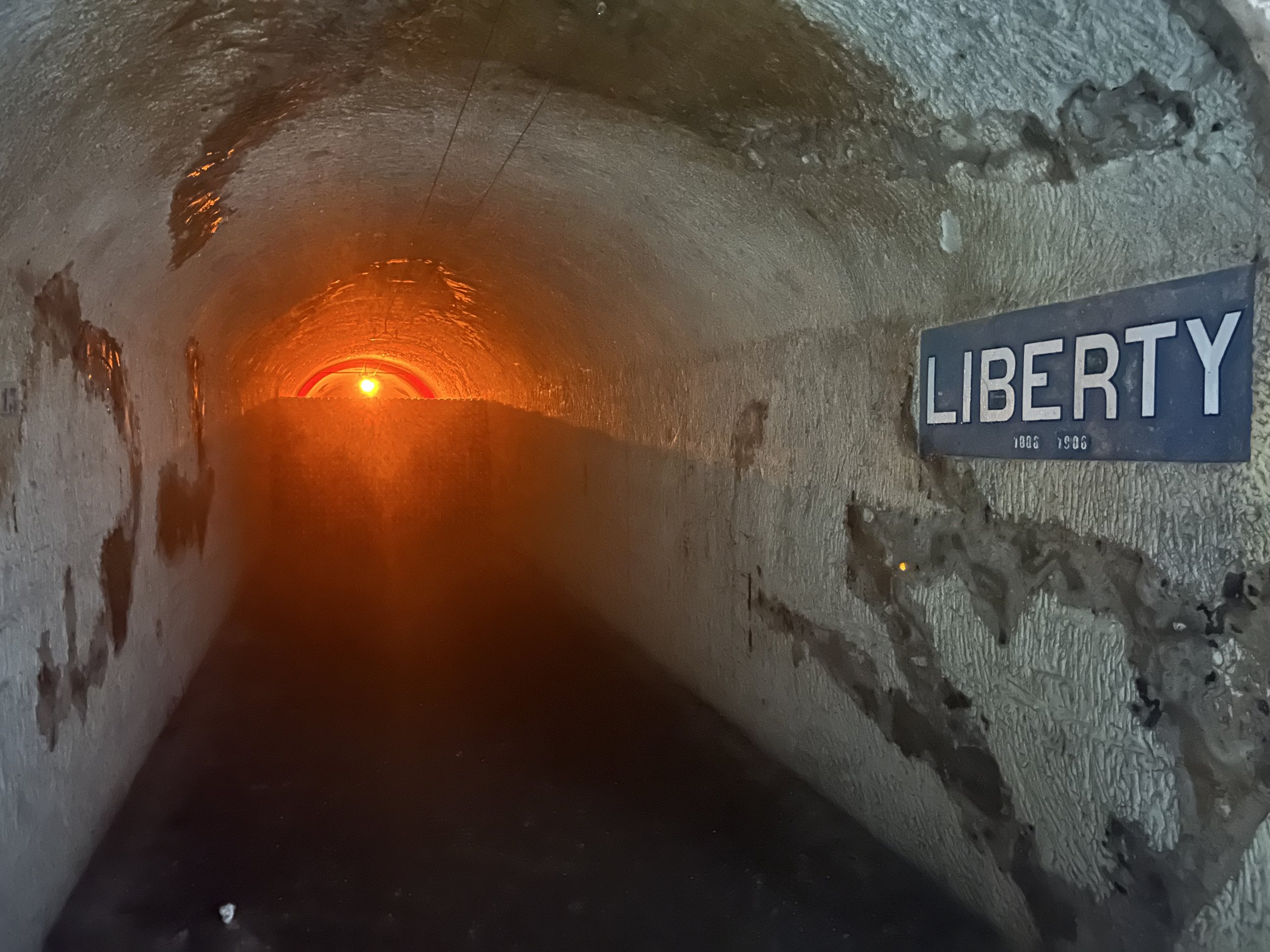 Inside a tunnel or underground passage with a sign that reads "Liberty" and a date range from 1906 to 1908, illuminated by an orange light at the far end.