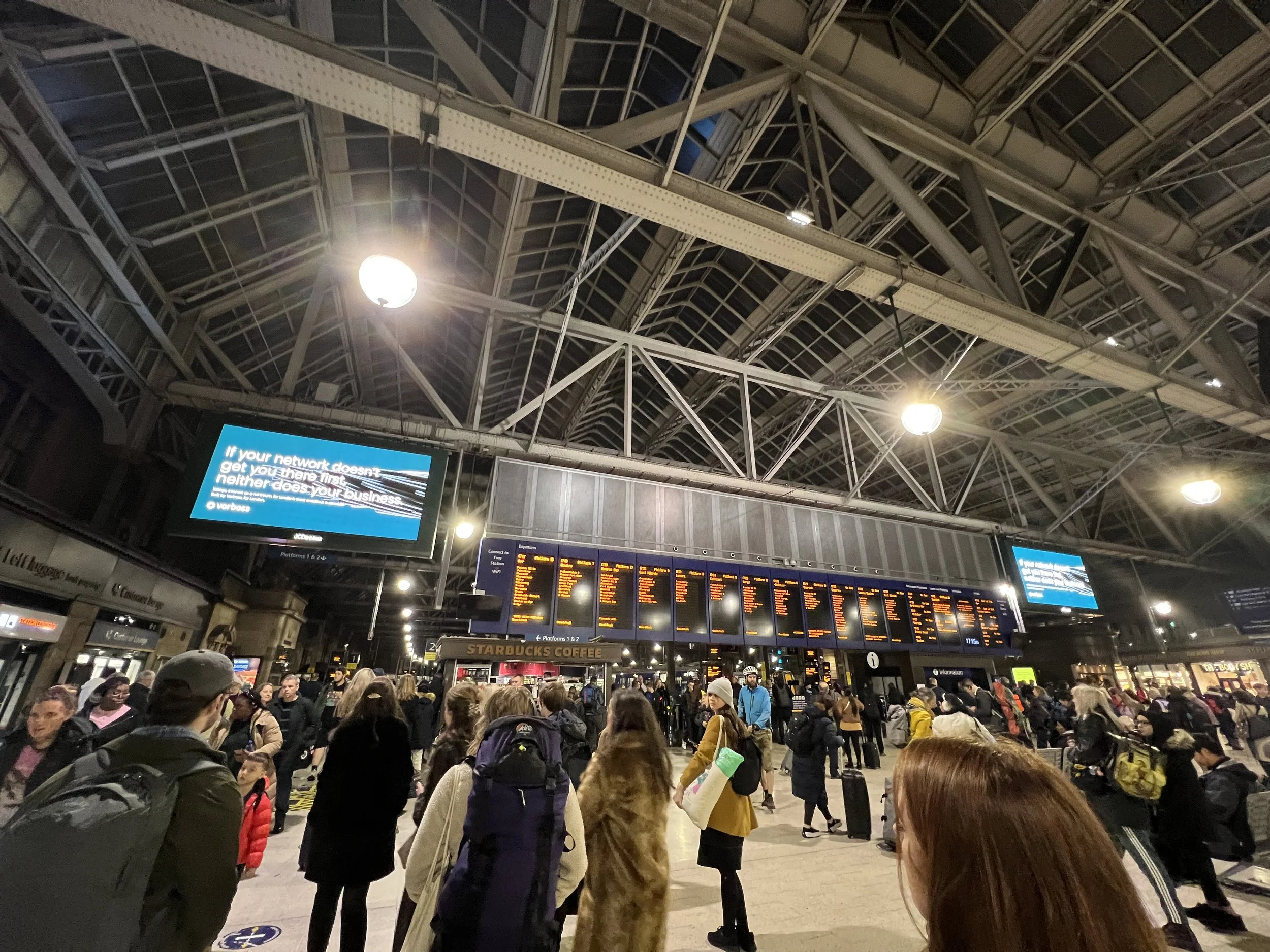 Crowd of people waiting at a train station, with digital departure screens and a Starbucks coffee shop visible, under a high arched metal roof.