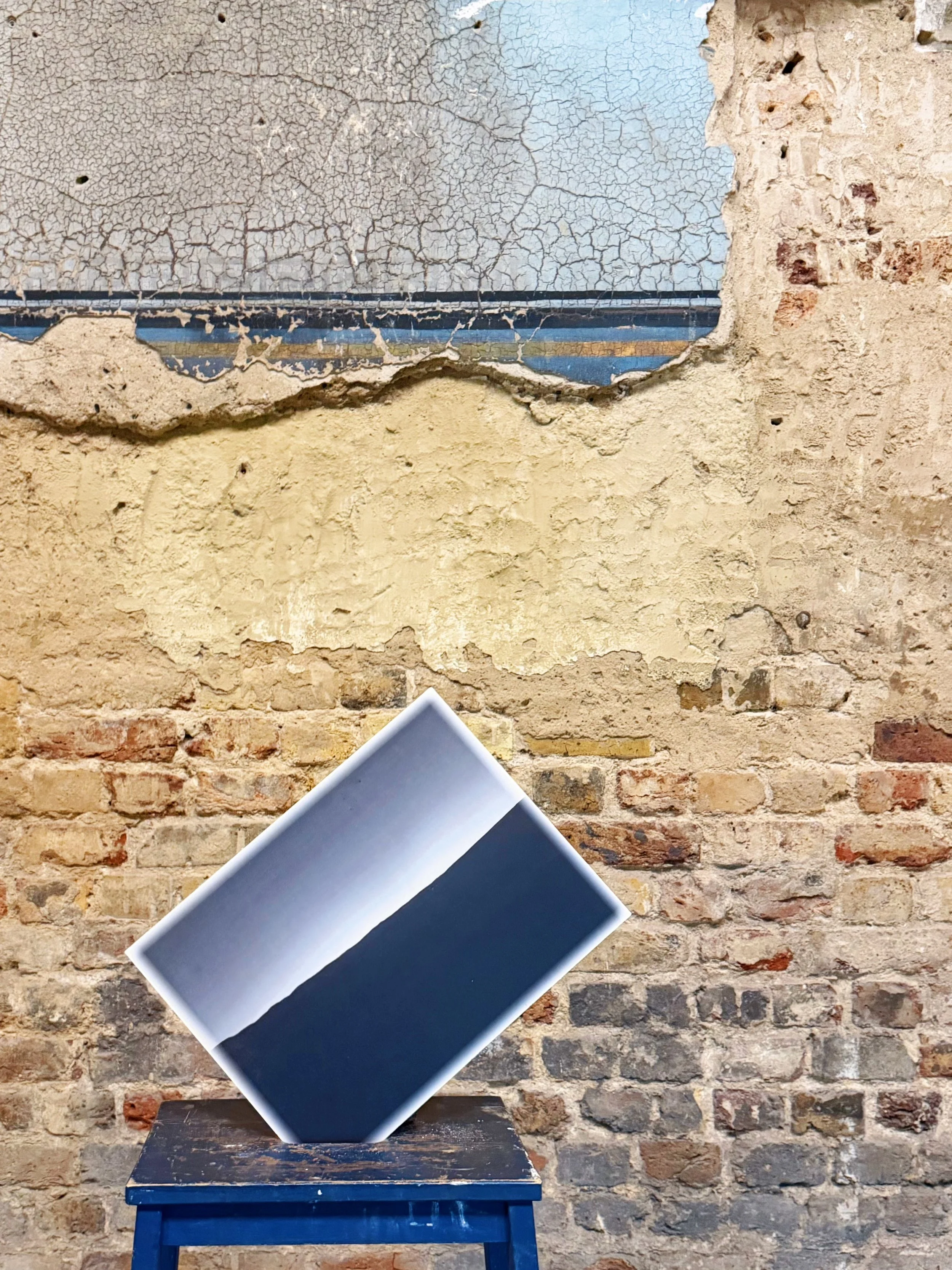A leather-bound book with a black cover leaning against a dark blue wooden stool, placed on a wooden surface in front of an exposed brick wall.