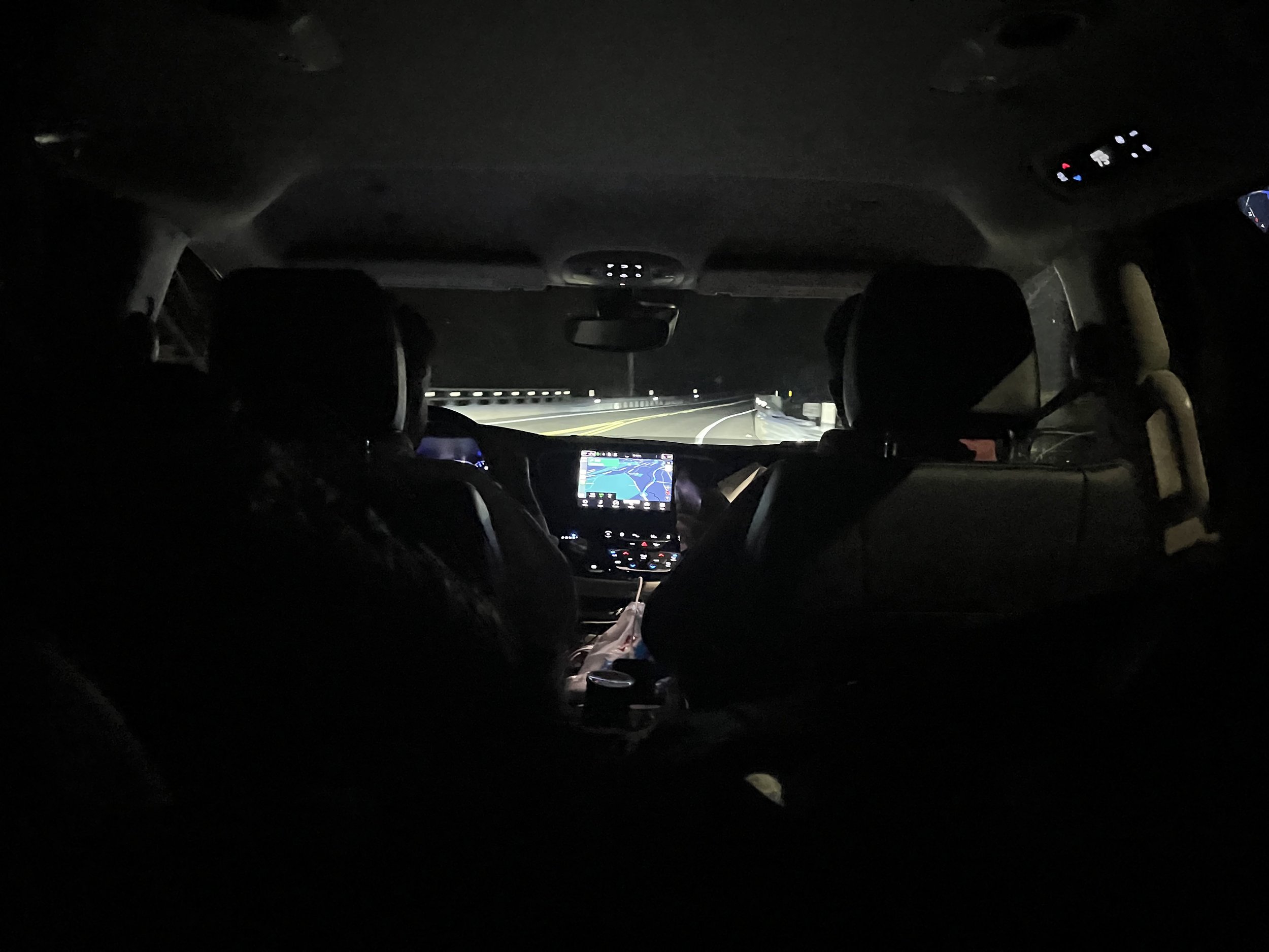 Inside a car at night, showing the dark interior, dashboard with GPS, and a view of the road ahead with streetlights.