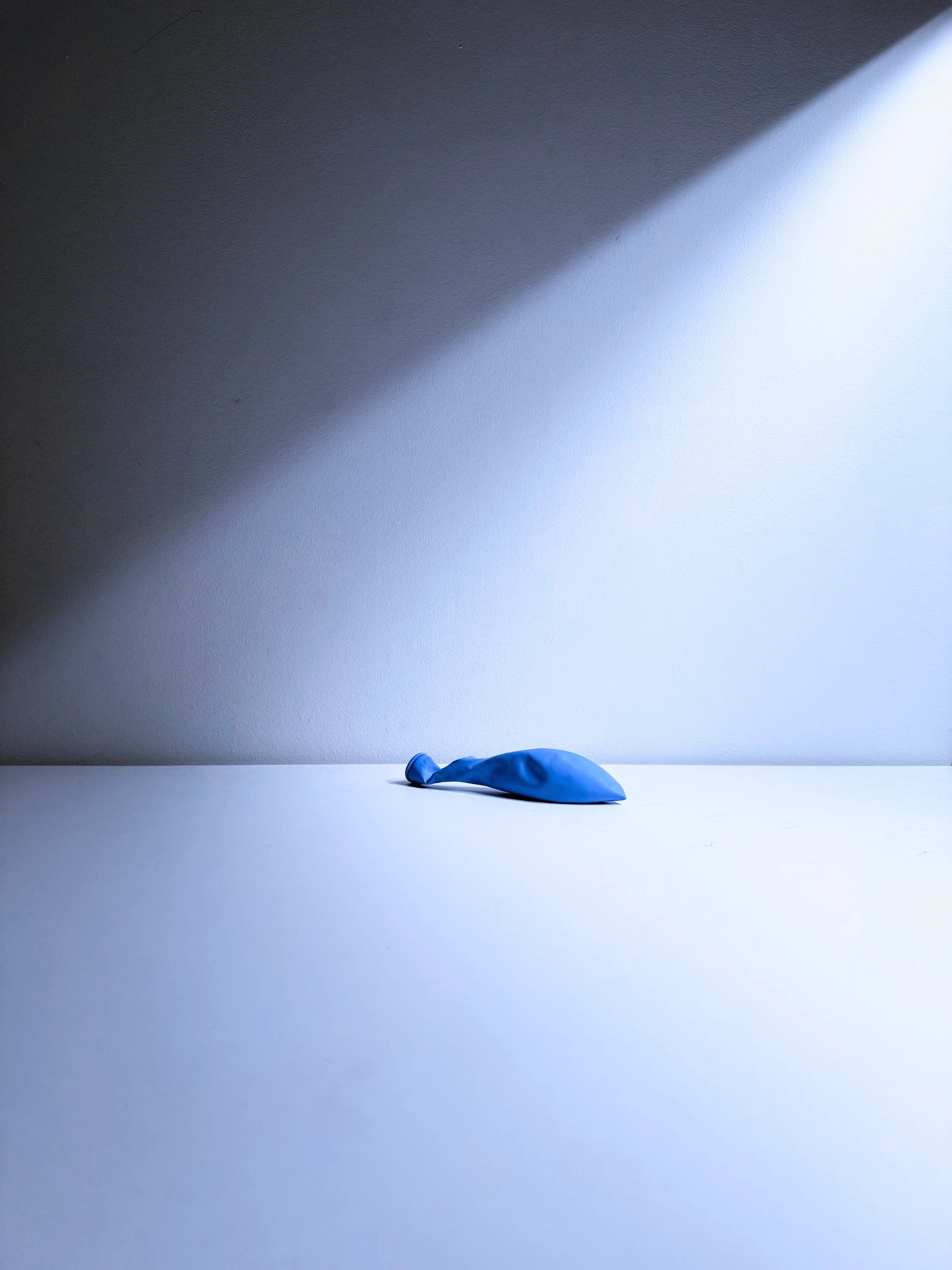 A blue balloon lying on a white surface with a shadow of light cast on the wall behind it.