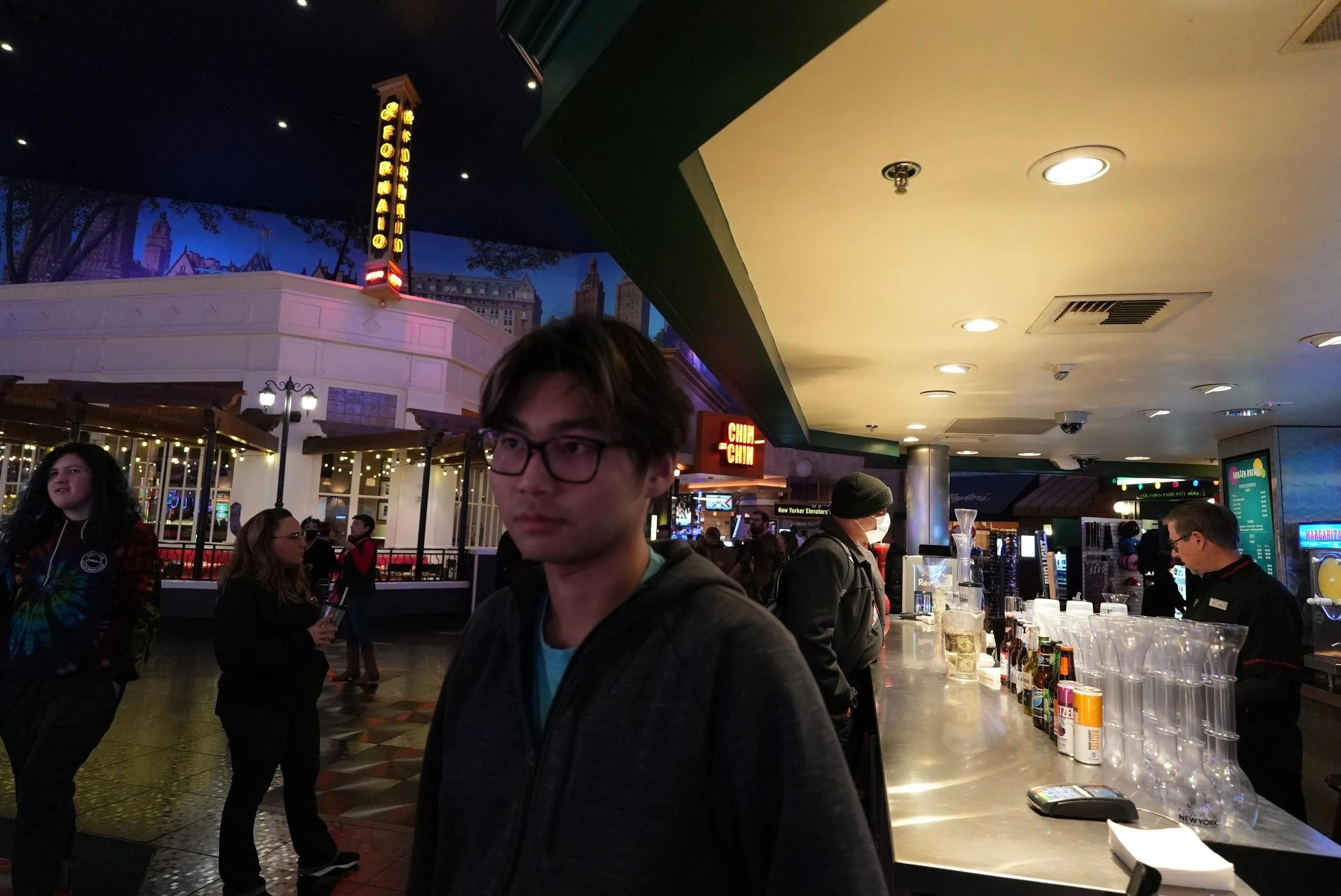 People standing at a bar in an indoor setting with a cityscape mural and neon signs in the background.