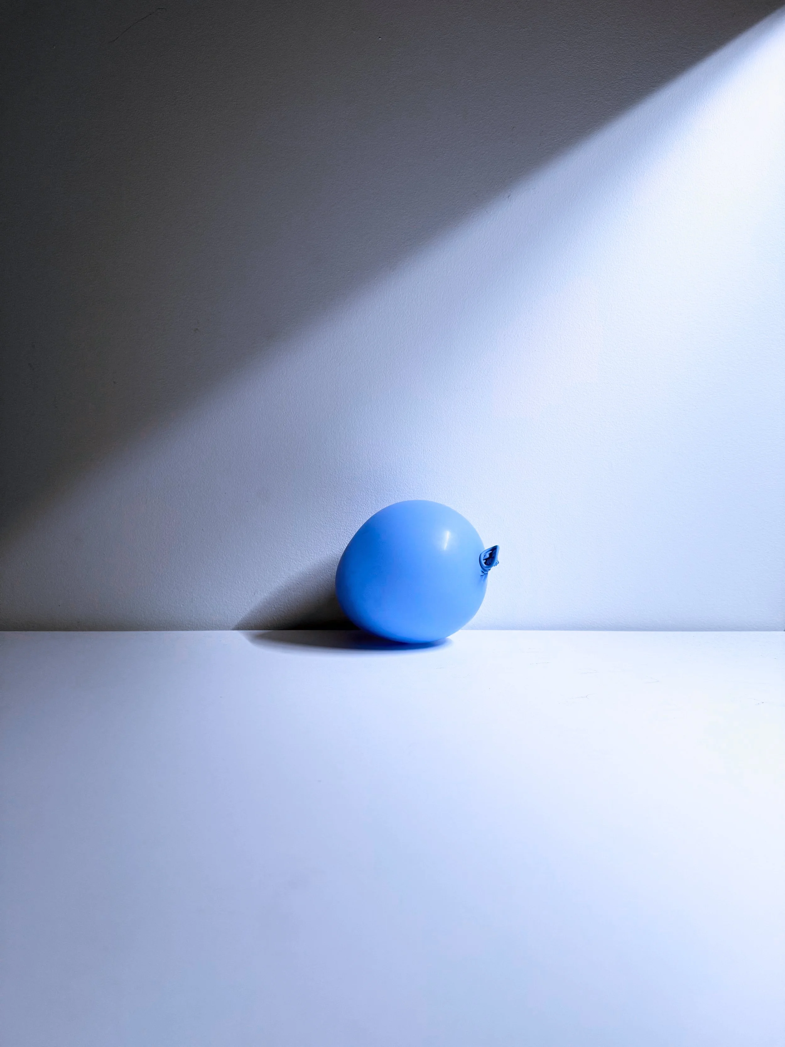 A single blue balloon with a small tear on its side, cast on a white surface with a shadow, against a two-tone wall with a dark upper half and a light lower half.