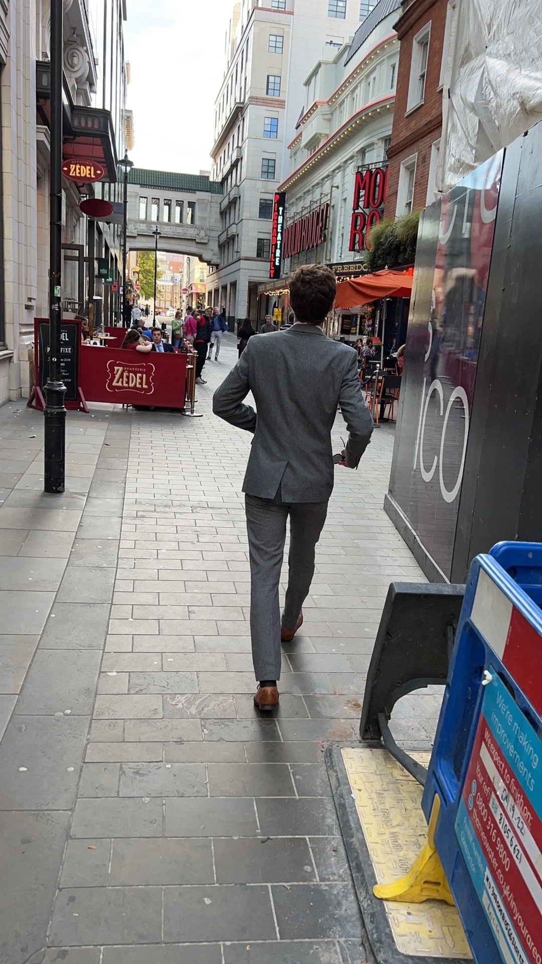 A man walking down a city sidewalk with various restaurants and stores on either side, including outdoor dining and signage.