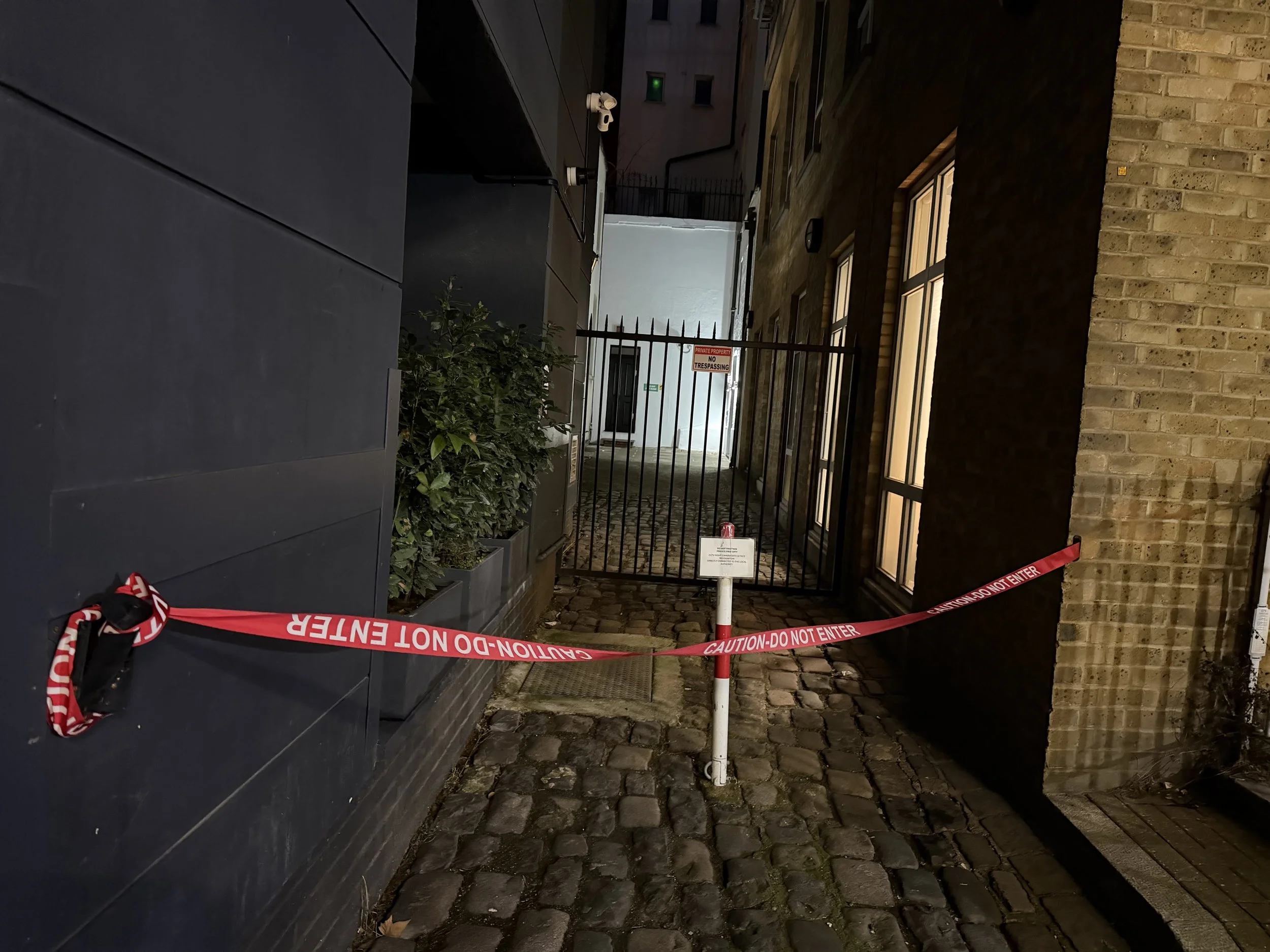 Nighttime alleyway with a black wall on the left, a brick building on the right, a gate with a 'No Trespassing' sign, a locked pole barrier with a 'Caution - Do Not Enter' tape, and cobblestone pavement.
