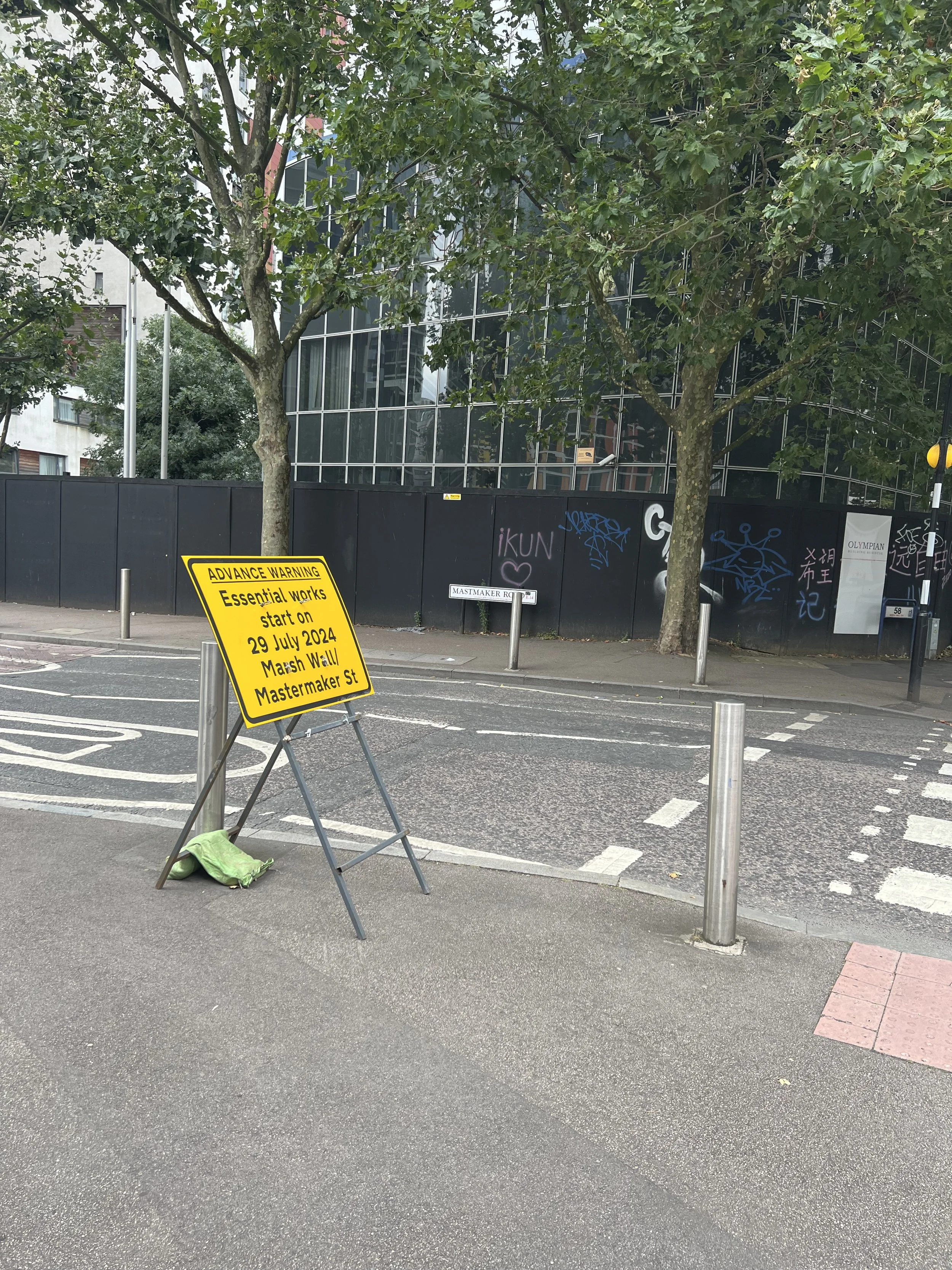 Street view with black construction fence, two trees, a yellow sign warning about essential work starting on July 29, 2024, at Marsh Wall, Mastermaker Street. There is graffiti on the fence and a street sign reading 'MASTMAKER ROW'. The scene include