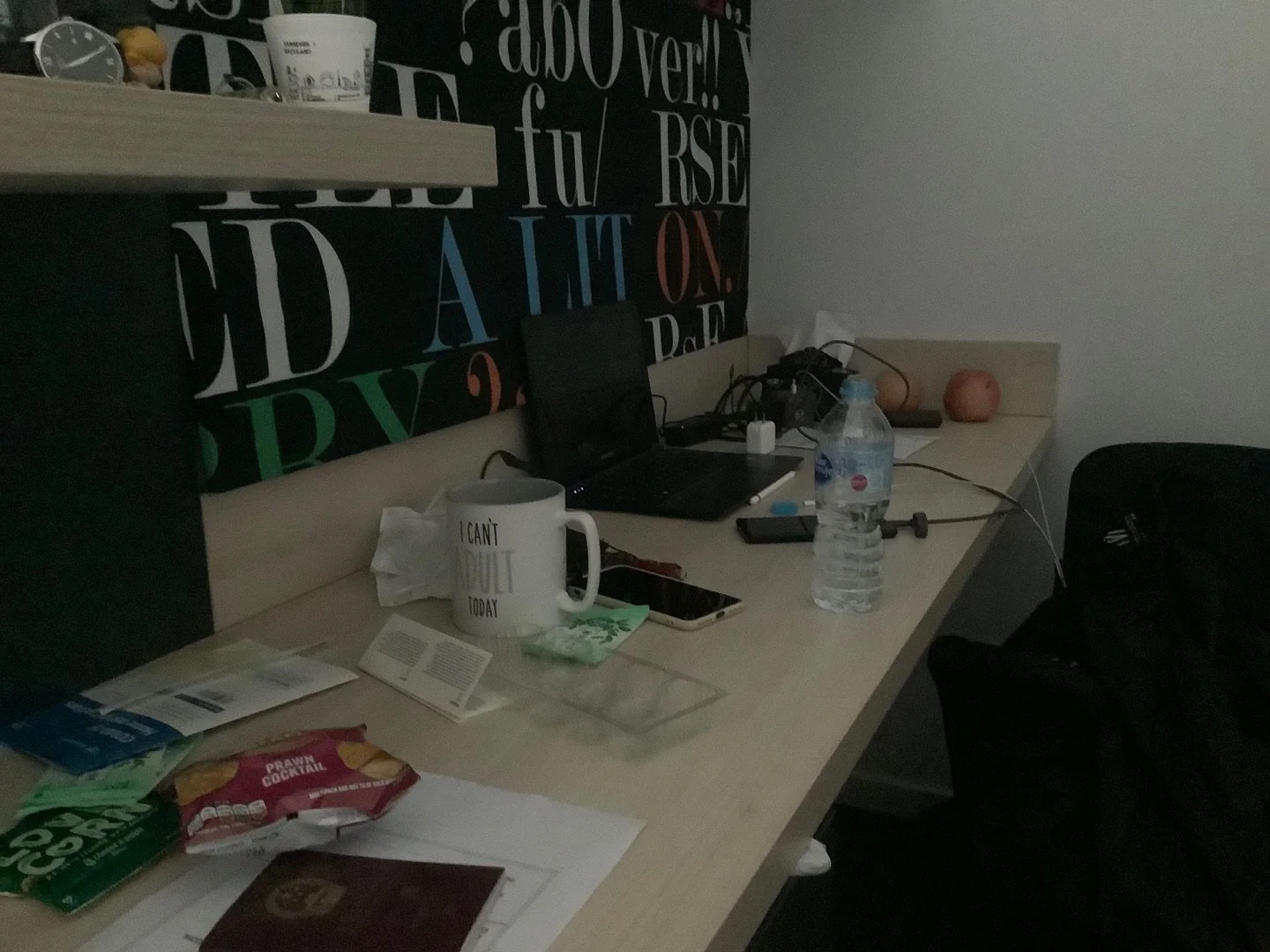 A cluttered desk with a large water bottle, a mug, a smartphone, a laptop, and various papers and snack packages. Two peaches are on the corner of the desk, and behind it is a black chair. The wall has colorful alphabet letters and words.