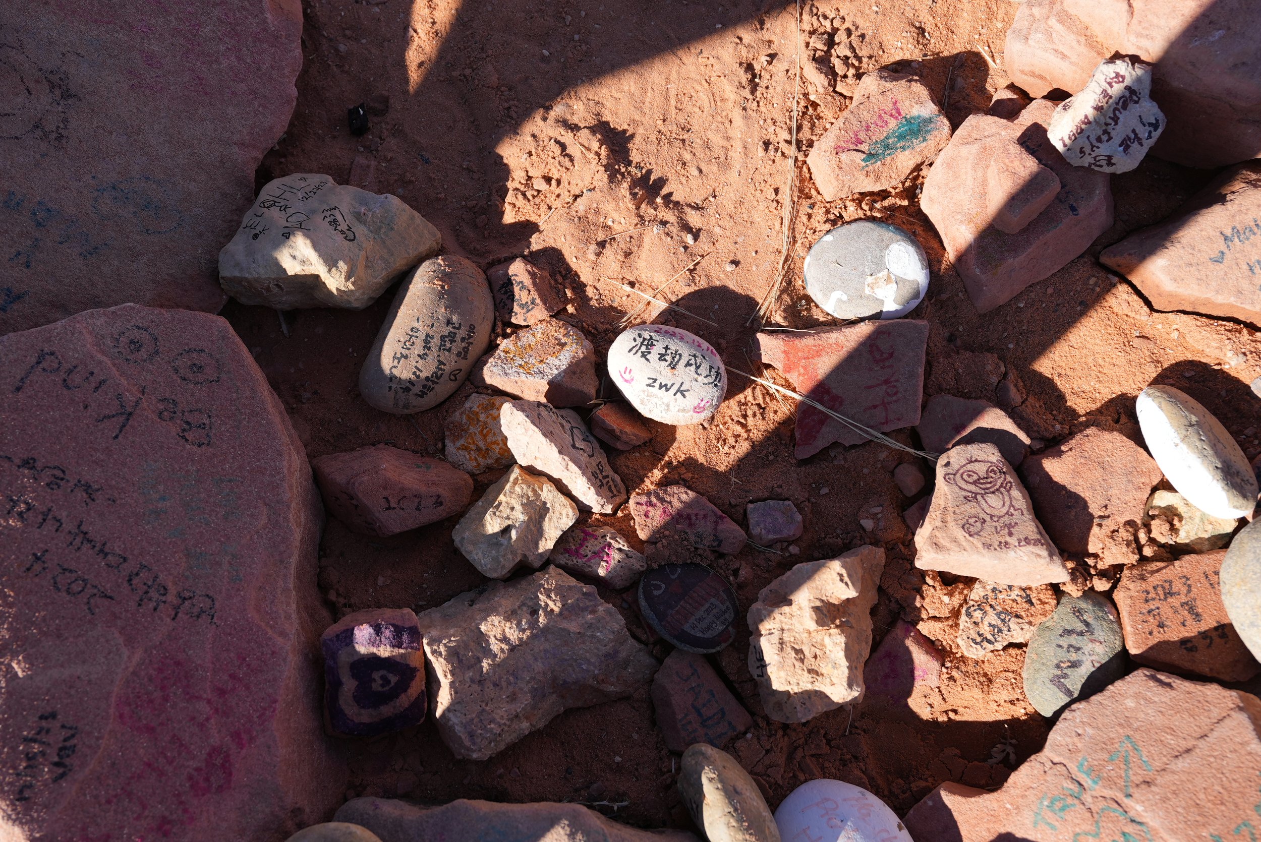 Rocks and stones with writings and drawings on them, placed on reddish dirt ground, some casting shadows.