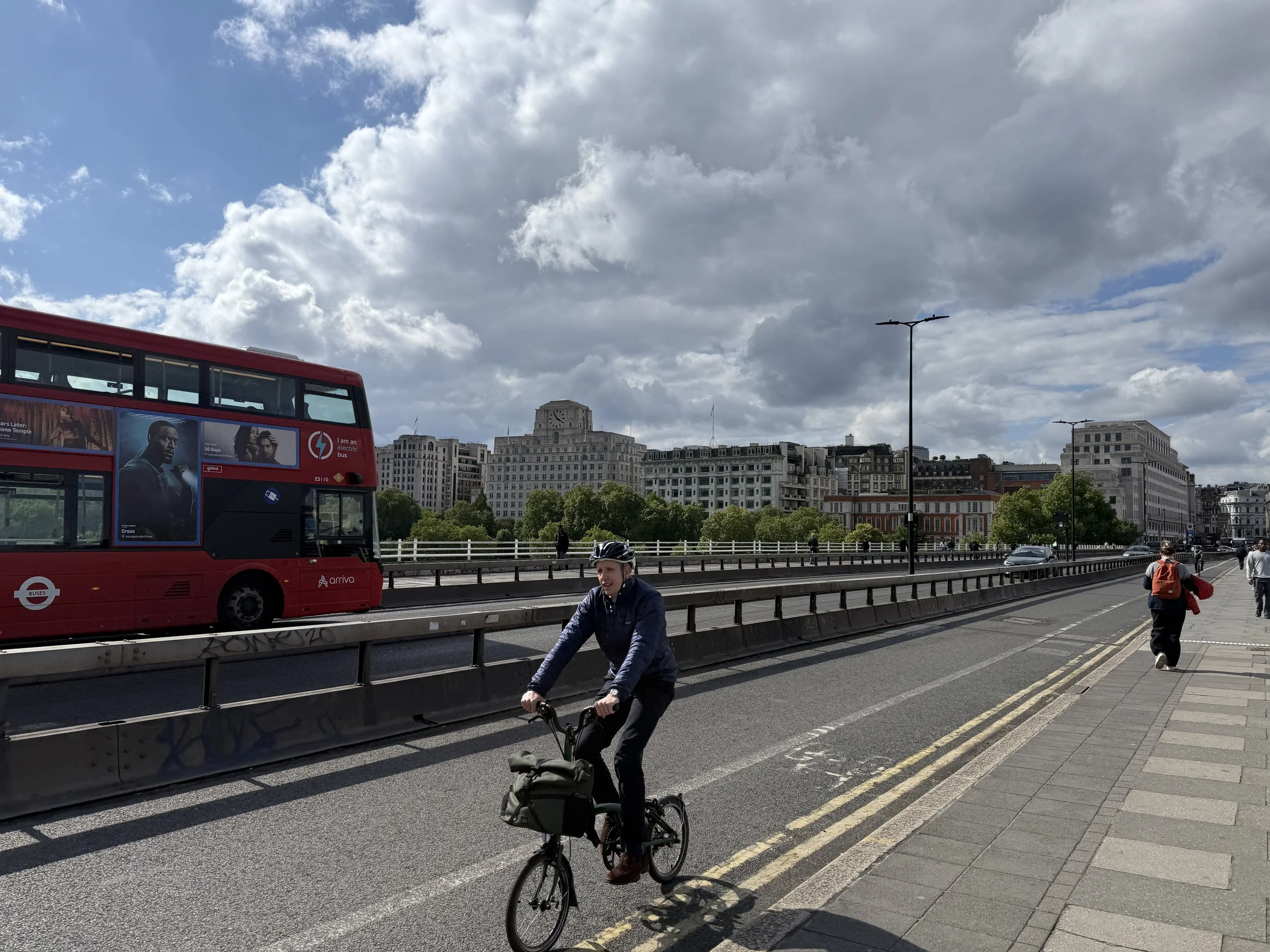 A city scene with a red double-decker bus, a person riding a bike, pedestrians, and historic buildings under a partly cloudy sky.