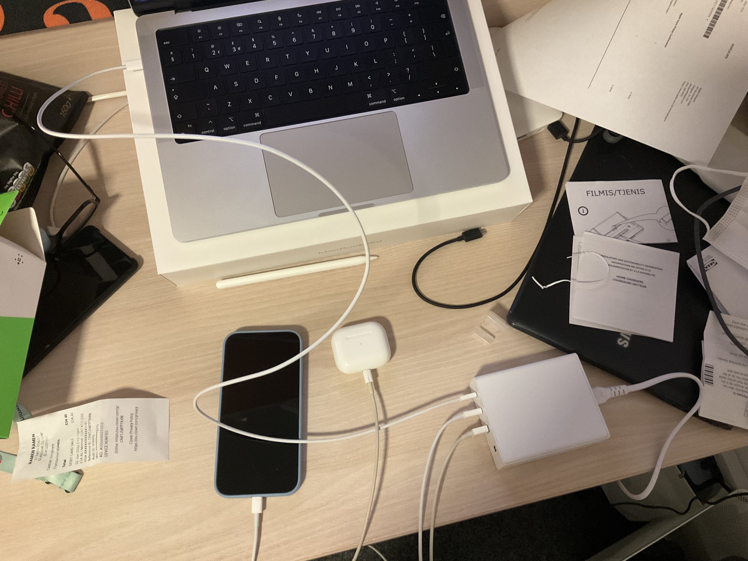 Desk with a MacBook, an iPhone, charger cables, papers, and other electronic devices scattered around.
