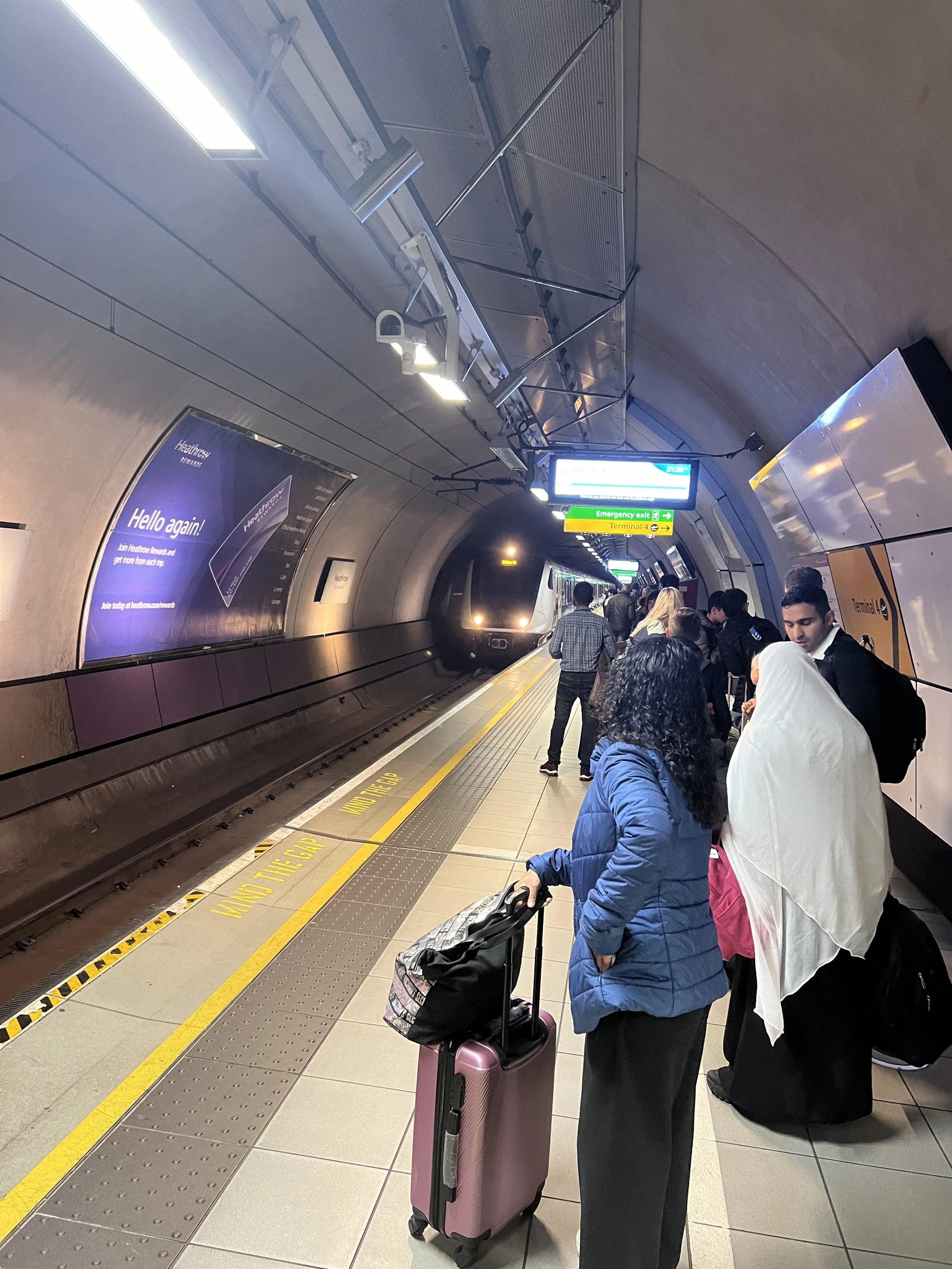 People waiting in line at an airport terminal, with some holding luggage and others with rolling suitcases, on a moving walkway, with digital signs overhead and advertisements on the side wall.