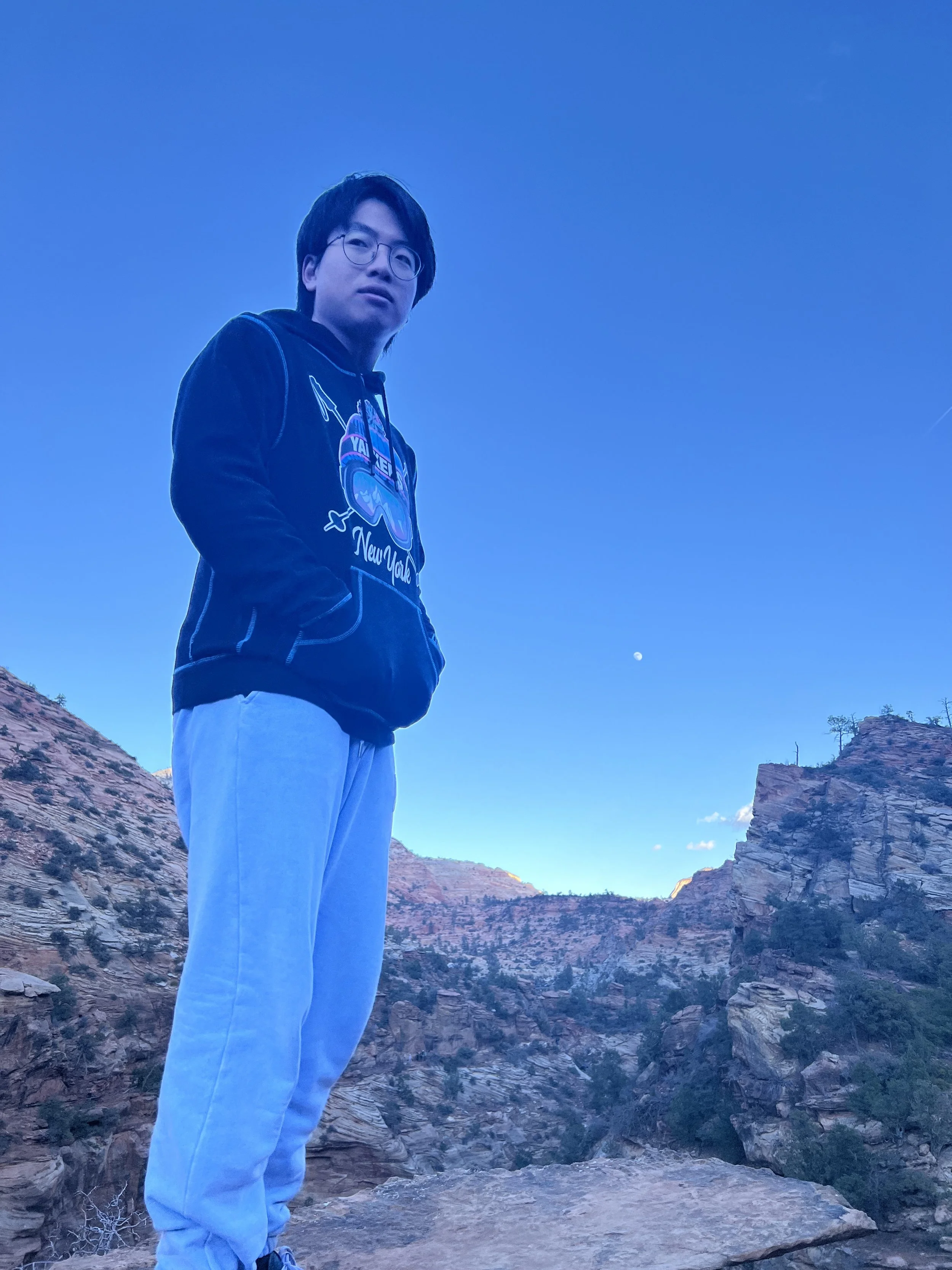A young man wearing glasses, a black hoodie with a colorful design, and light blue pants, stands on rocky terrain with canyon cliffs and a clear blue sky in the background. The moon is visible in the sky.