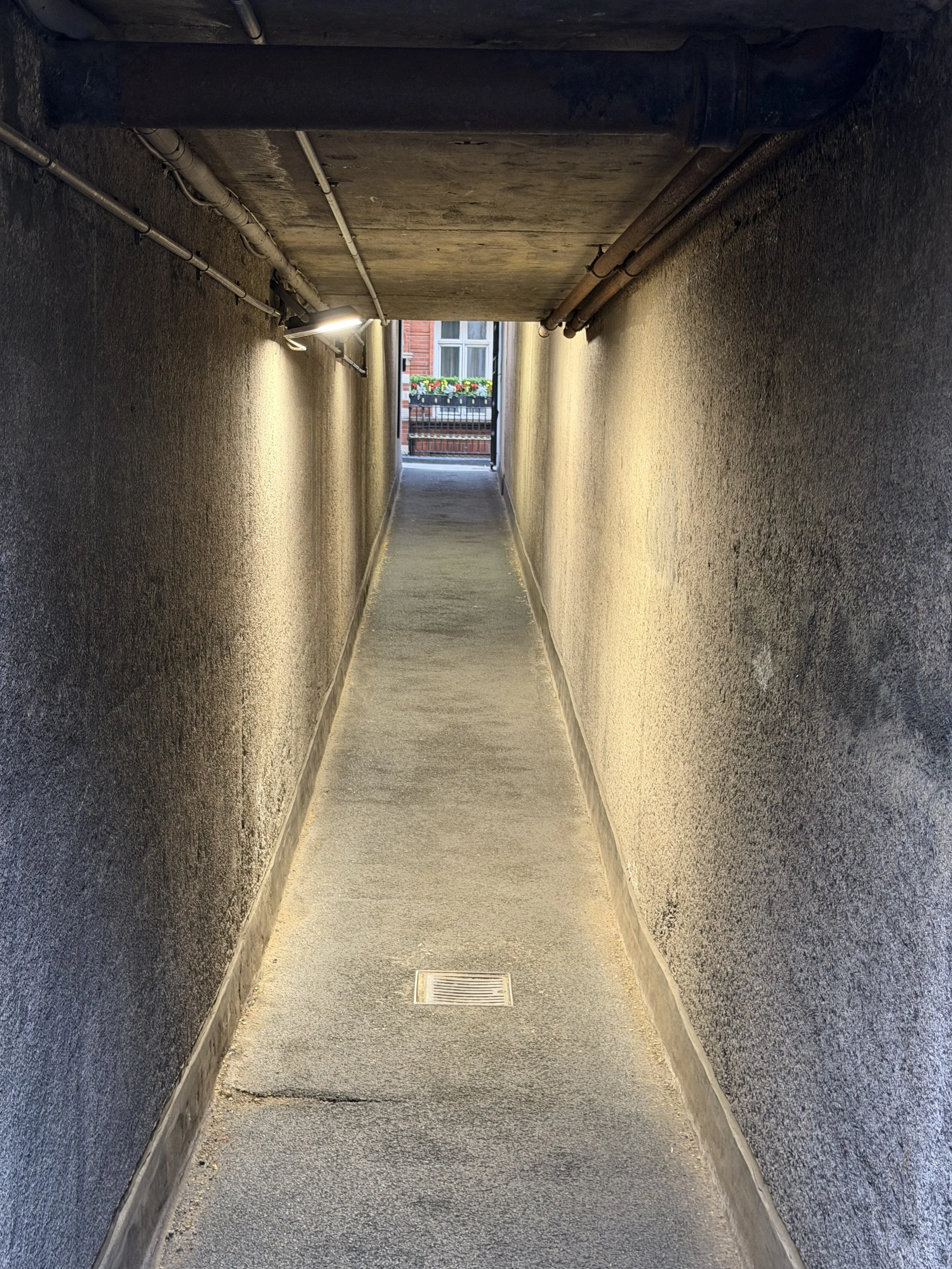 A narrow underground walkway with textured beige walls, a drainage grate on the floor, and a view of a brick building with a balcony and flower boxes at the end.