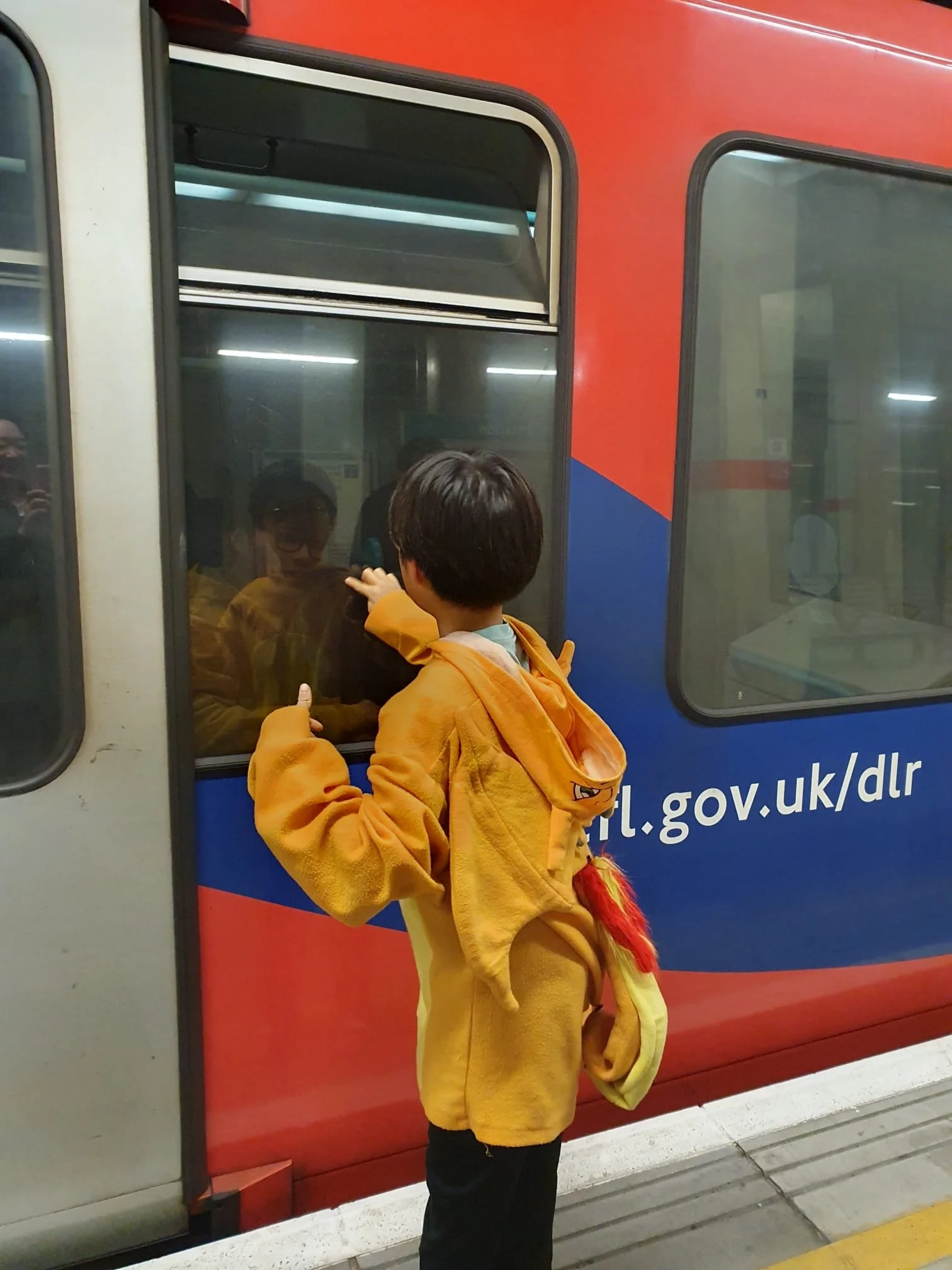 A young boy in a yellow jacket stands on a subway platform looking at a train, with his reflection visible in a window of the train.